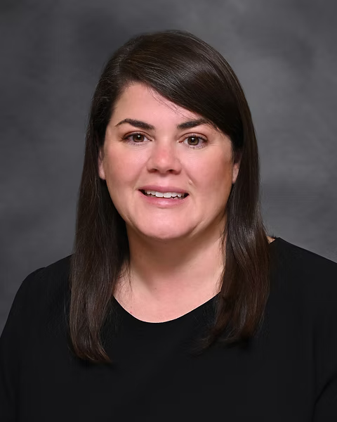 A professional headshot of a woman with dark brown hair and fair skin wearing a black top, smiling against a gray background.