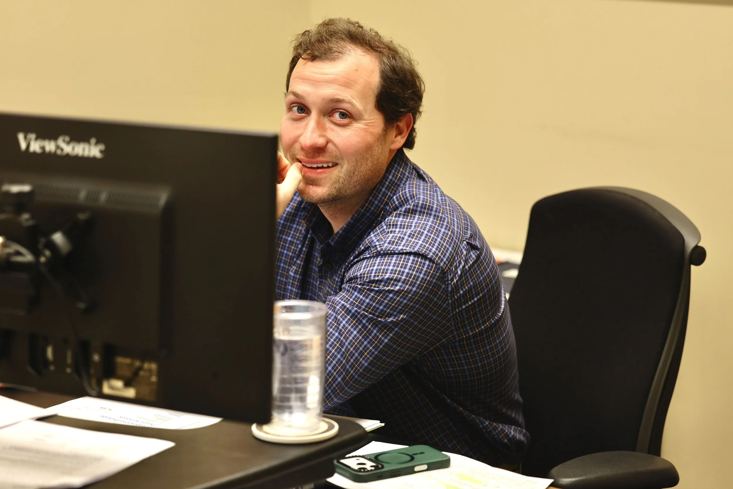 A man with dark hair and a beard, wearing a blue checkered shirt, sitting at a desk in front of a computer monitor, smiling and looking at the camera. There is a transparent water glass, some papers, and a green phone on the desk.