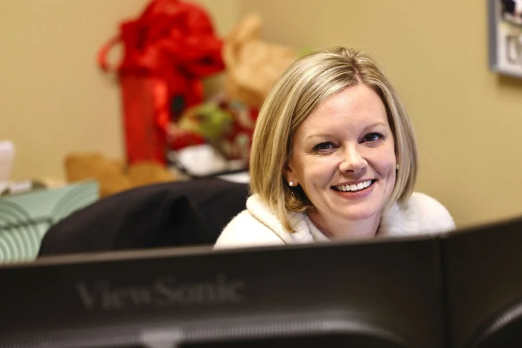 A woman with blonde hair smiling while sitting at a desk with a computer monitor in front of her.