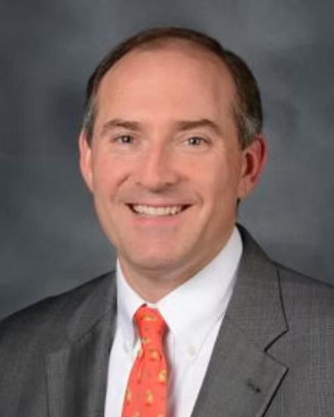 Professional headshot of a man in a gray suit with a white shirt and a red patterned tie, smiling against a dark background.