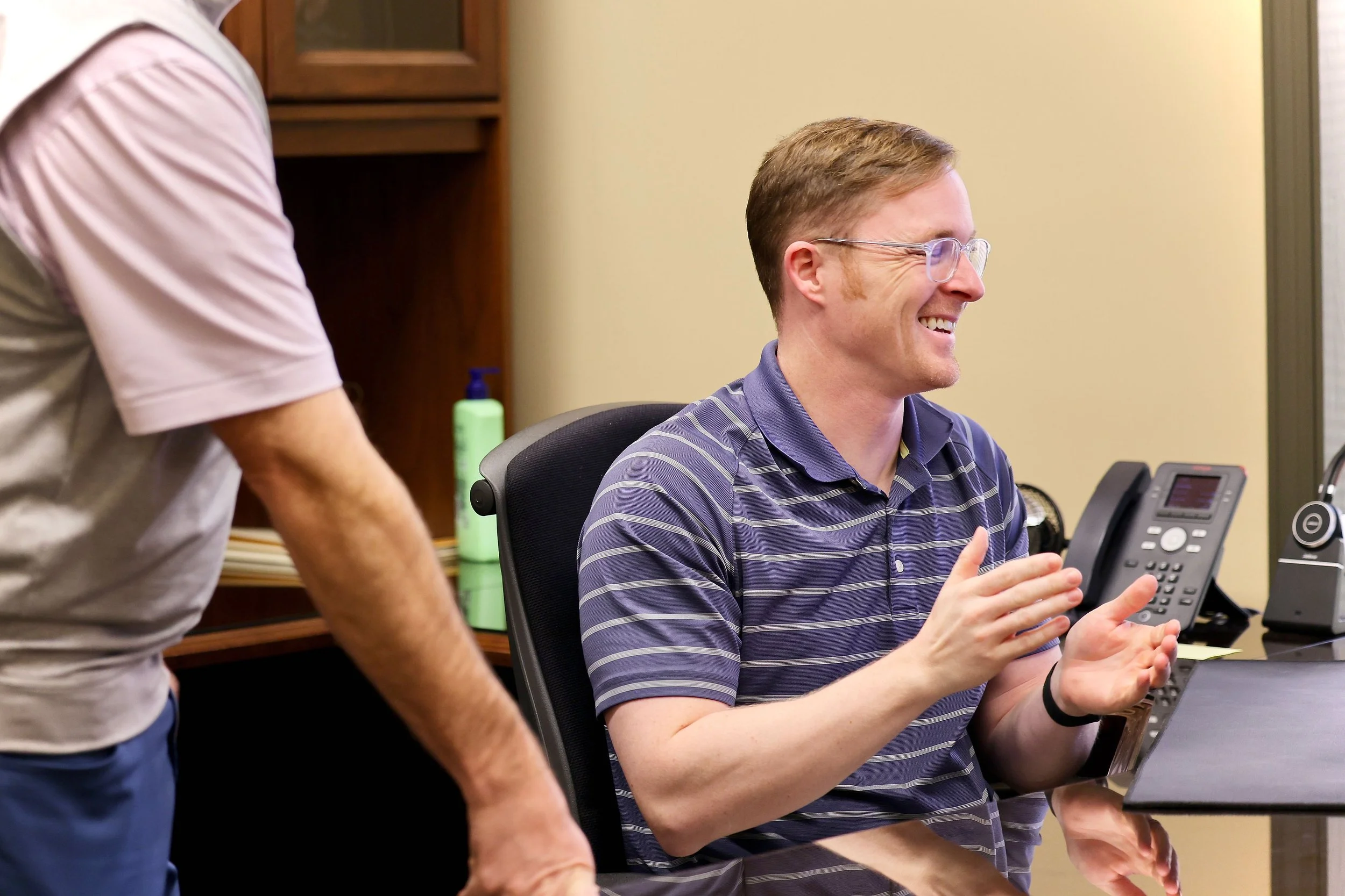 A man smiling and clapping while sitting at a desk in an office, with another person standing nearby. The man has glasses and is wearing a blue striped polo shirt.
