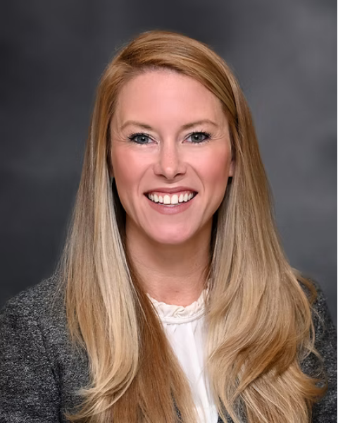 A professional headshot of a woman with long blonde hair, smiling, wearing a white blouse and gray blazer against a dark gray background.
