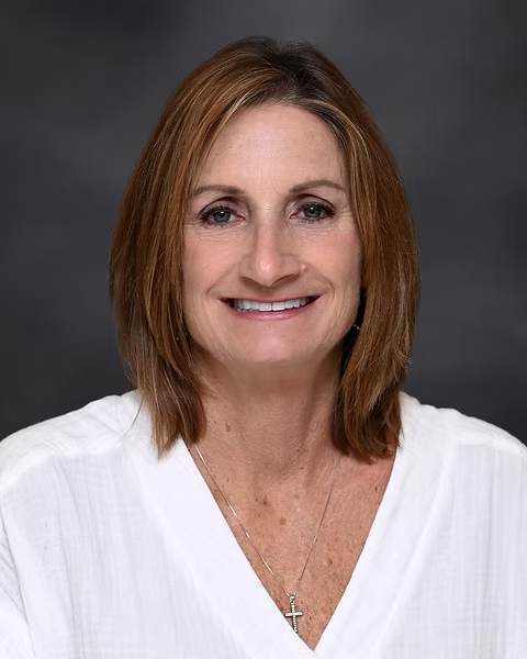 A professional headshot of a woman with shoulder-length light brown hair, smiling, wearing a white top and a cross necklace against a dark gray background.