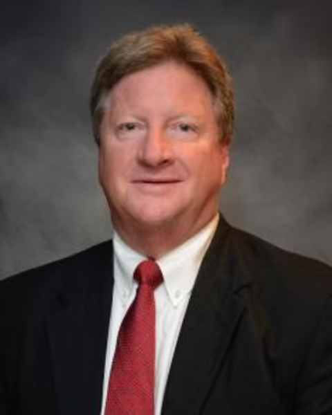 A professional headshot of a man with light brown hair, wearing a dark suit, white shirt, and red tie, against a dark gray background.