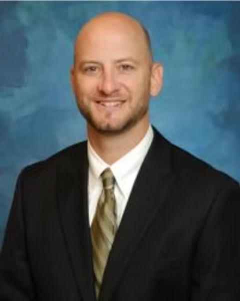 A professional headshot of a of a man in a suit with a tie against a blue background.