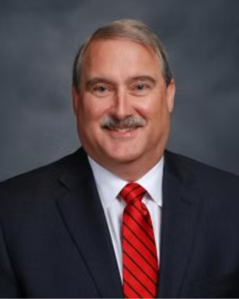 A professional headshot of a man wearing a black suit, white shirt, and red striped tie, smiling in front of a gray studio background.