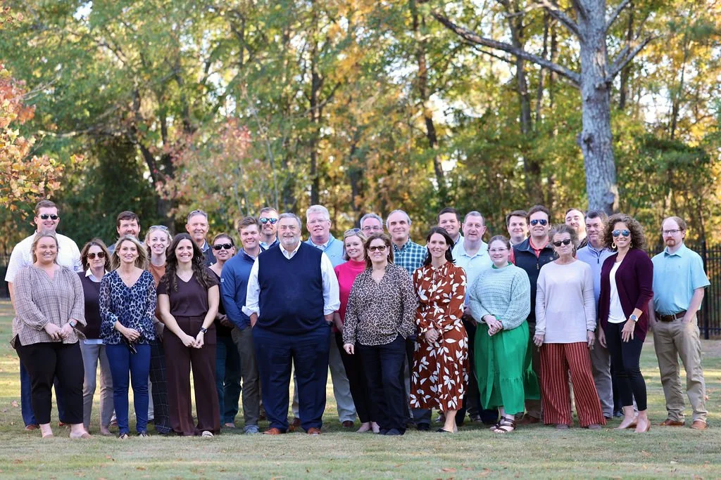 A group of people outdoors in a park during autumn, standing on grass with trees and colorful leaves in the background.