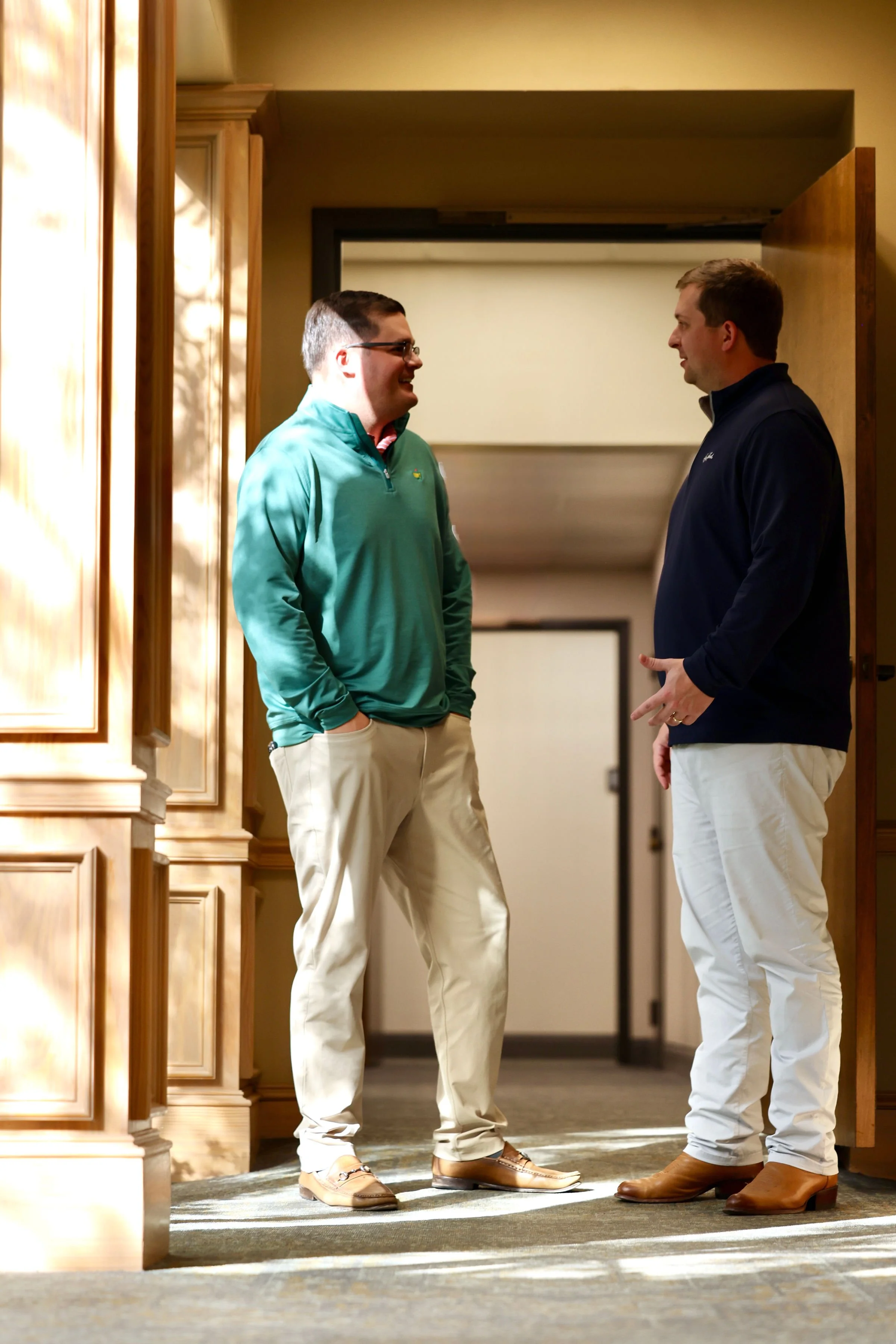 Two men standing and talking in a hallway with wooden cabinet and door, both wearing casual attire with khakis and boots, engaged in a conversation and smiling.