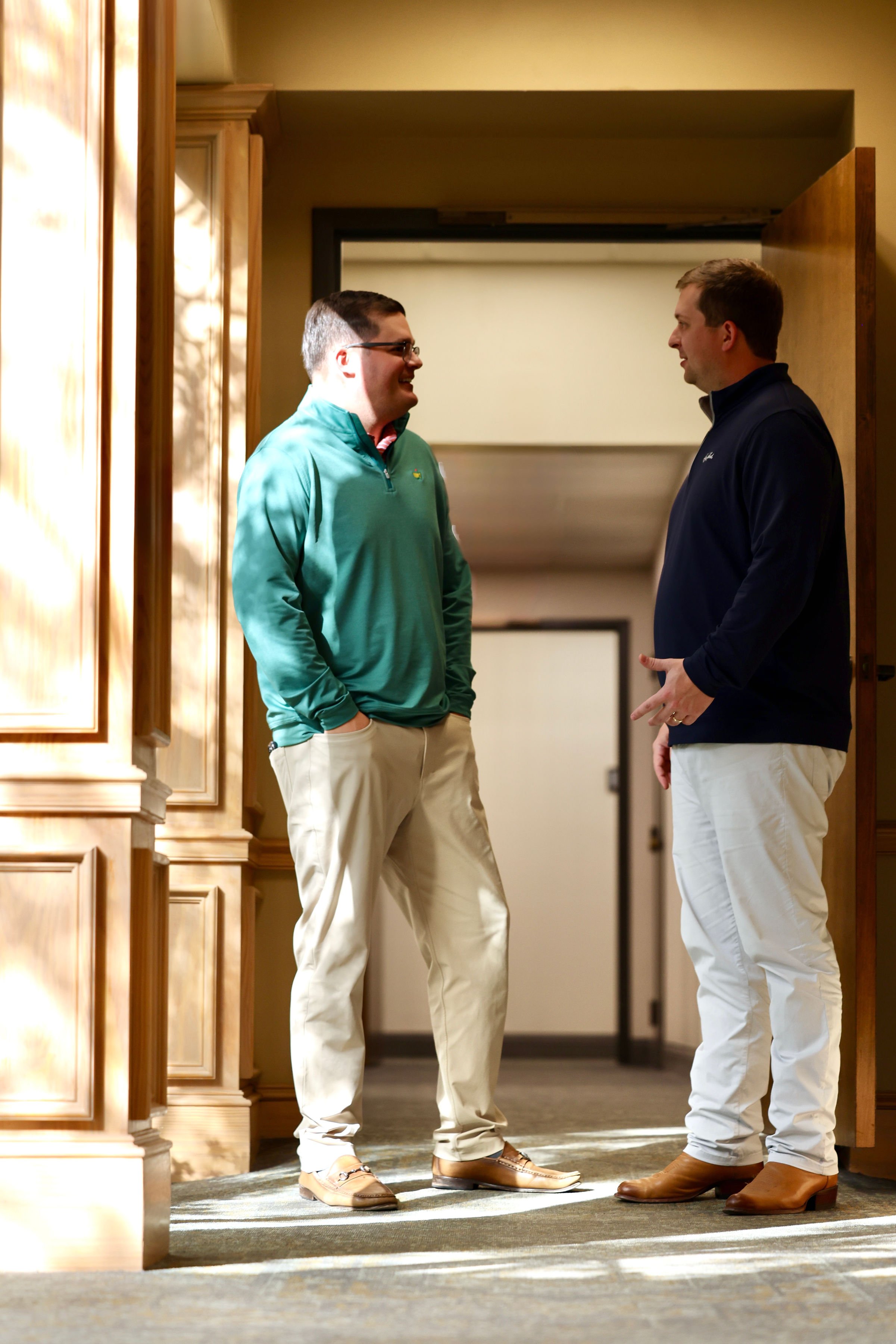 Two men are standing and talking indoors near a wooden wall, with sunlight casting shadows on the wall. One man is wearing a green jacket with beige pants, and the other is in a navy jacket with beige pants. They appear to be having a conversation.