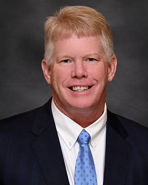 A professional headshot of a smiling man wearing a dark suit, white shirt, and a light blue tie, against a dark gray background.