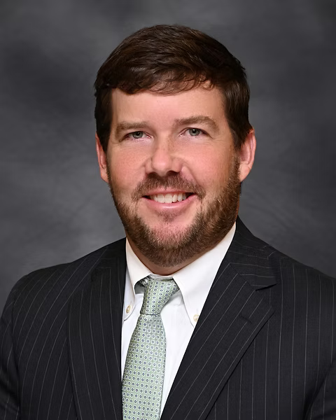 A professional headshot of a man with brown hair, beard, wearing a dark pinstriped suit, white shirt, and light-colored patterned tie, smiling against a dark background.