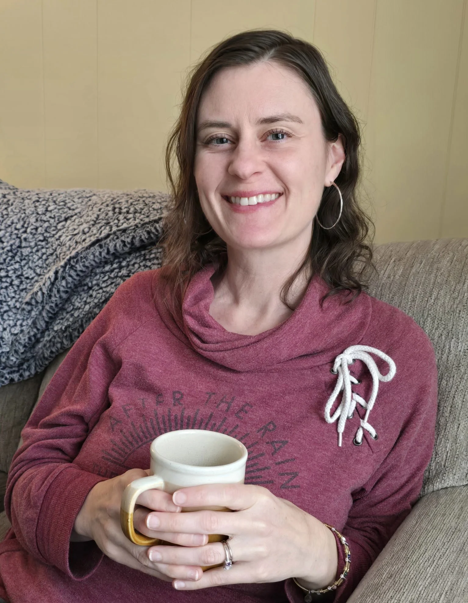A woman with brown wavy hair smiling and holding a beige cup, sitting on a beige couch with a gray blanket in the background, wearing a maroon hoodie with a white drawstring.