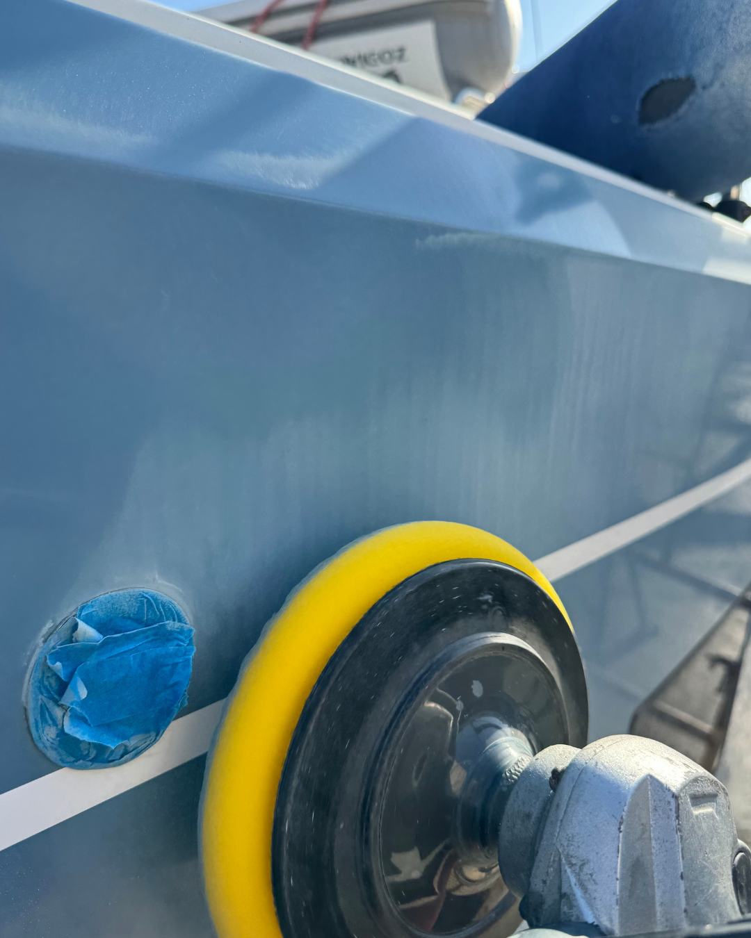 Close-up of a power buffer or polisher with a yellow pad, working on a gray surface, possibly a vehicle, with a small piece of blue masking tape nearby. pulir casco barco , alicante, taller náutico, el campello