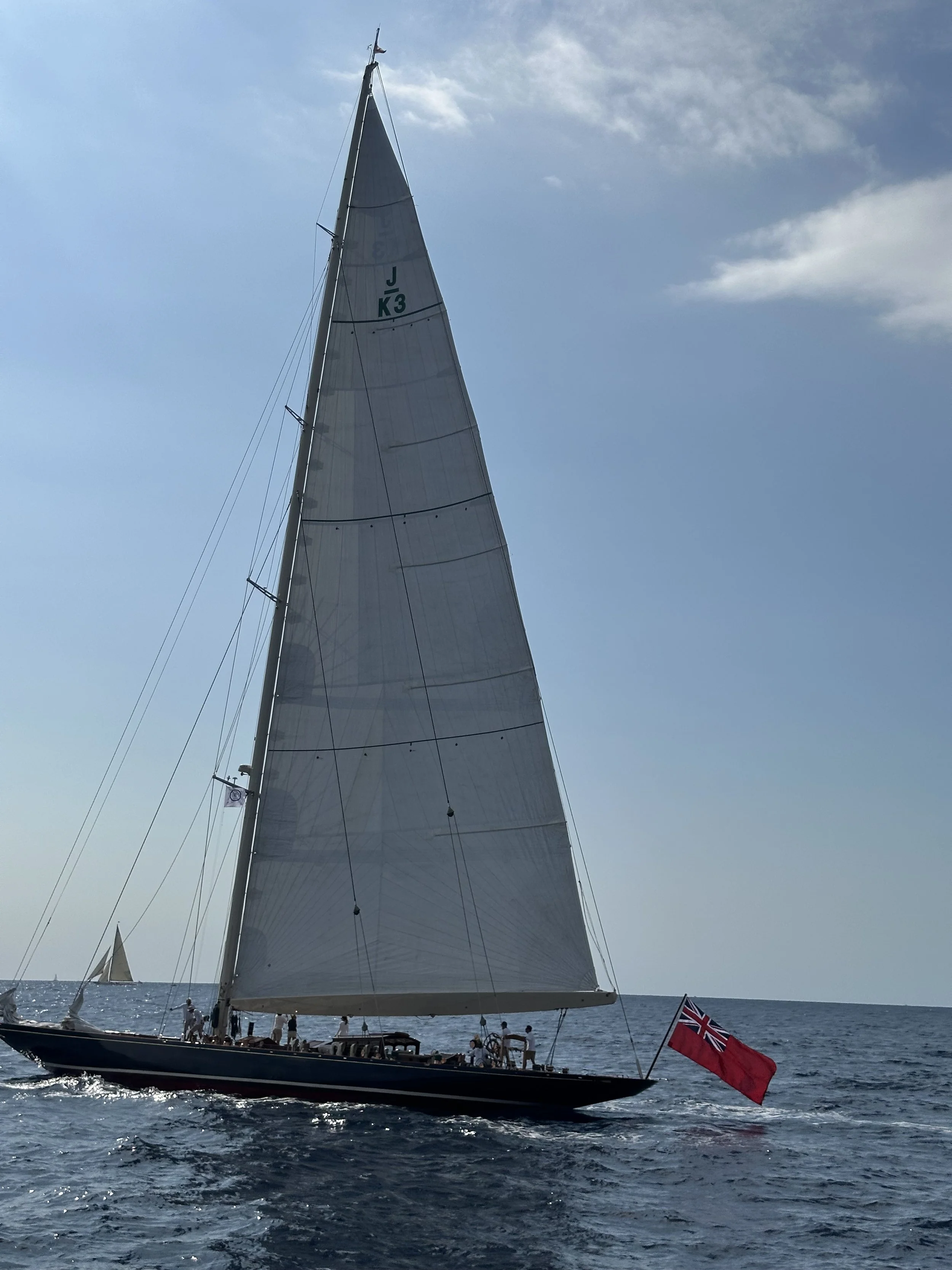 Classic racing yacht with tall white sails and a British red ensign competing in the Régates Royales in the Bay of Cannes