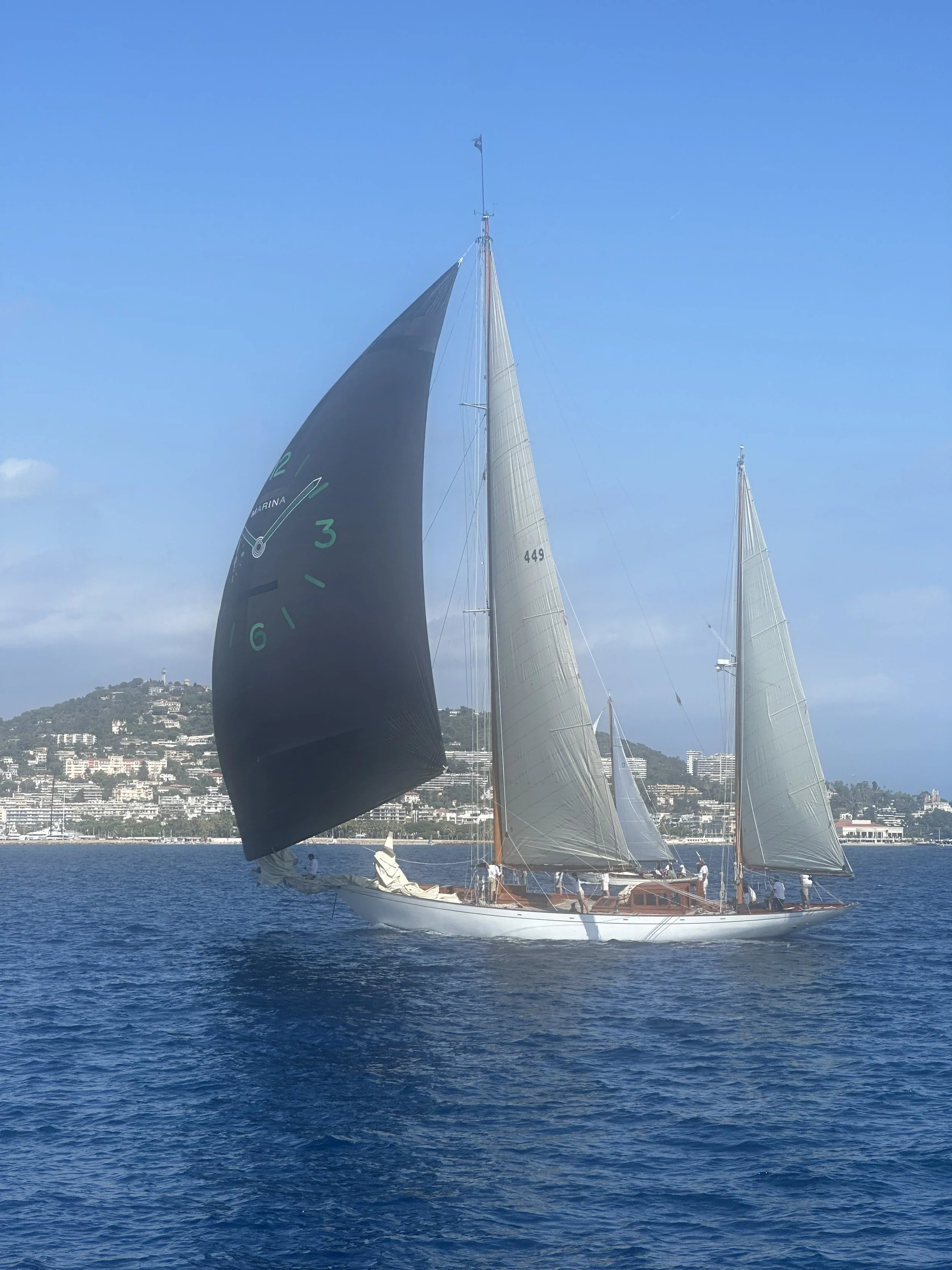 Classic yacht number 449 flying a black spinnaker as it races past the Cannes coastline during the Régates Royales
