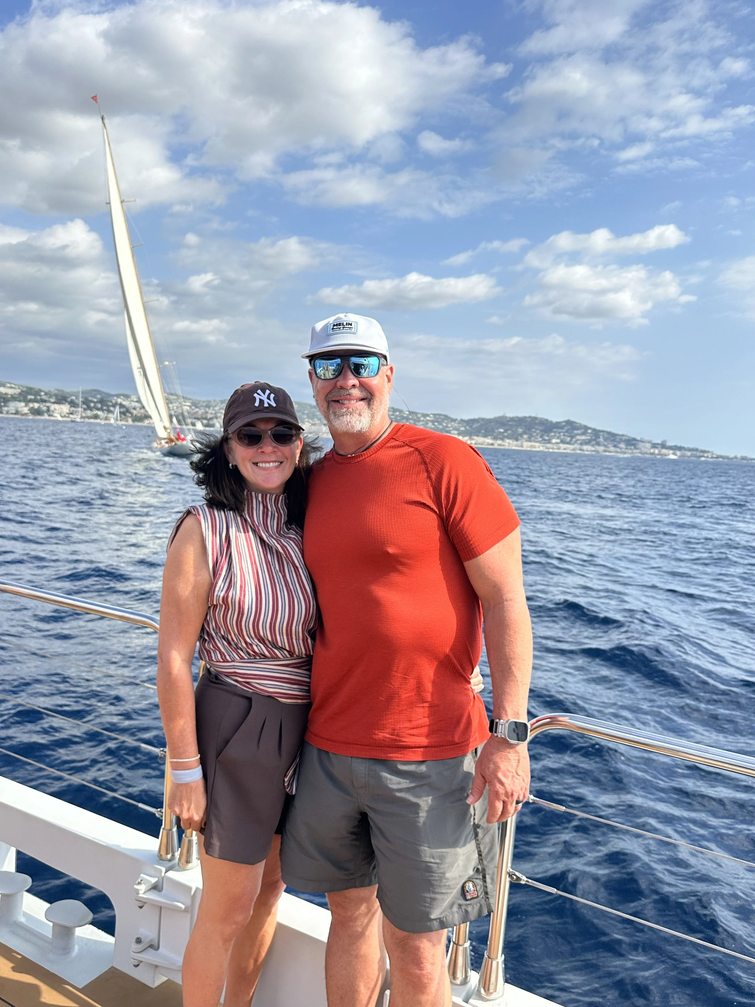 Couple on the deck of a sailing catamaran during the Régates Royales in Cannes, with classic yachts and the French Riviera coastline behind them