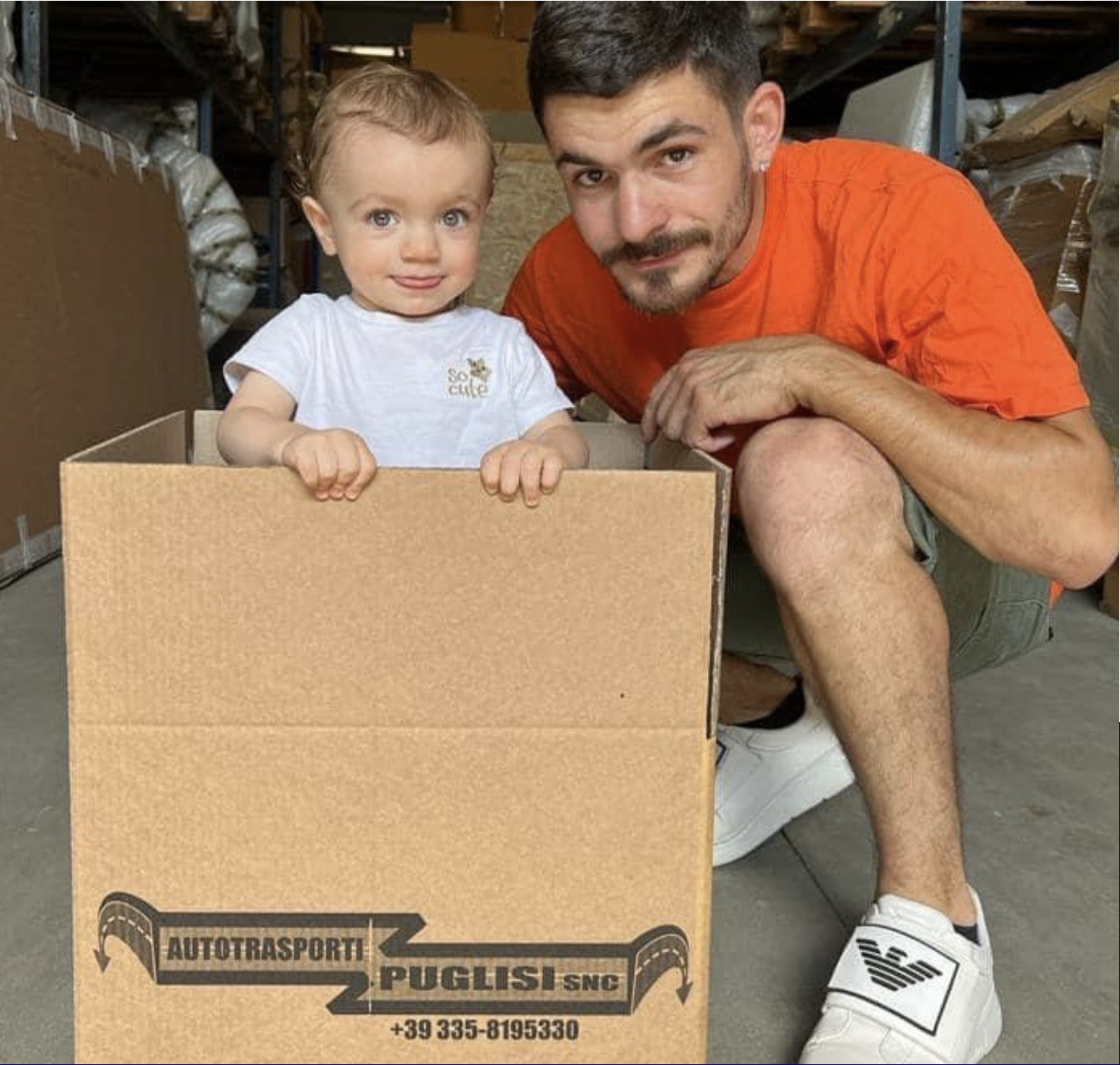 Un bambino e un uomo sorridente sono accovacciati vicino a una scatola di cartone in un magazzino. La scatola ha il logo 'Autotrasporti Puglisi SNC' e un numero di telefono. Entrambi guardano verso la fotocamera, con il bambino che si aggrappa al bordo della scatola e l'uomo che si appoggia con un braccio sulla sua gamba.