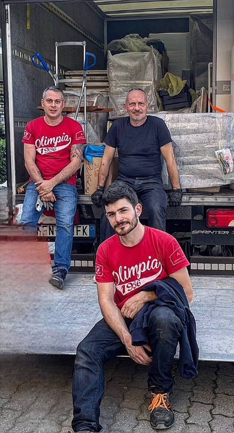 Tre uomini sorridenti davanti a un camion carico di scatole e materiali di imballaggio, uno seduto sul portellone del camion, gli altri in piedi dietro.