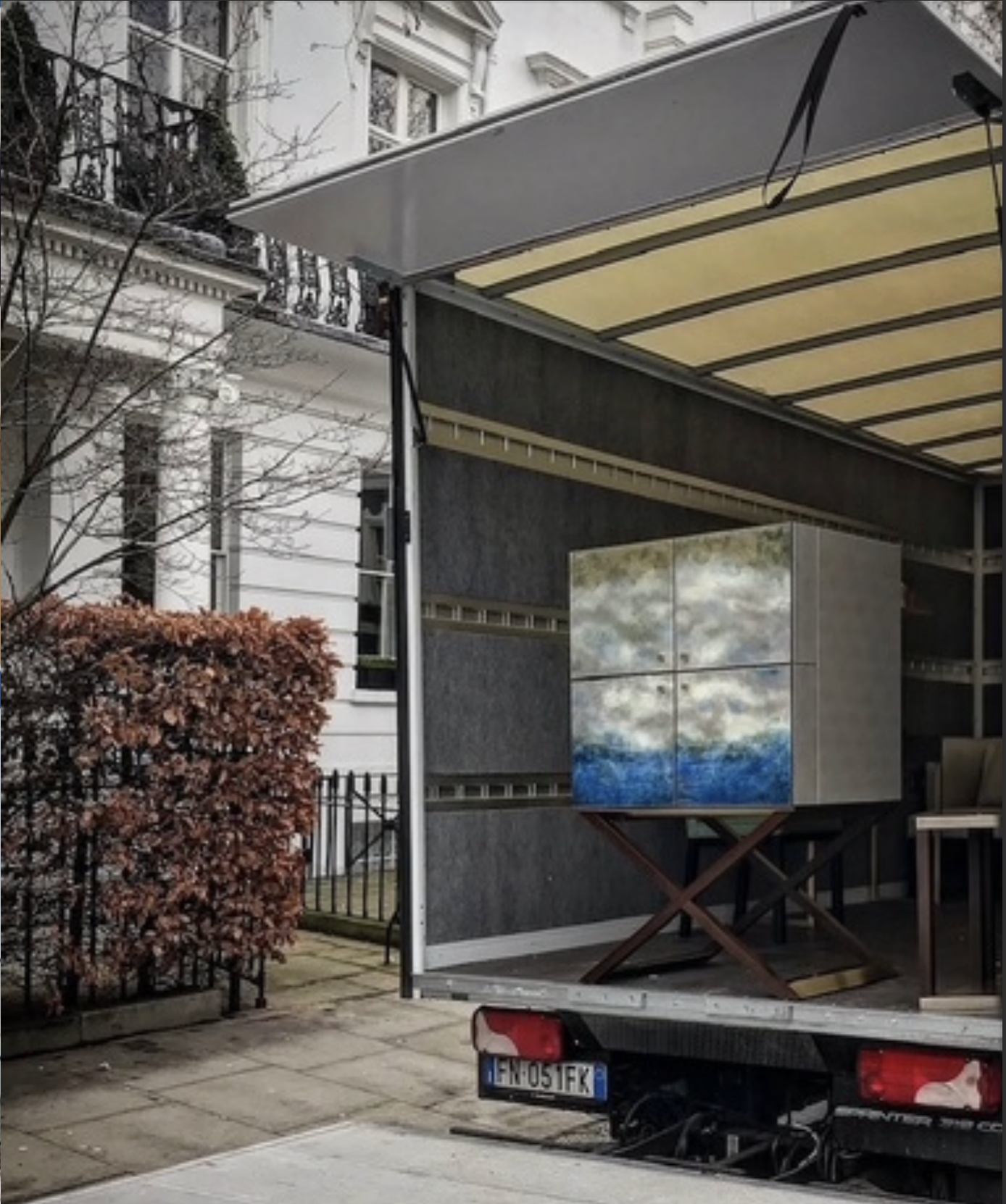 Un camion con un contenitore artistico con dipinti di nuvole e mare, parcheggiato davanti a edifici residenziali con balconi e una siepe.