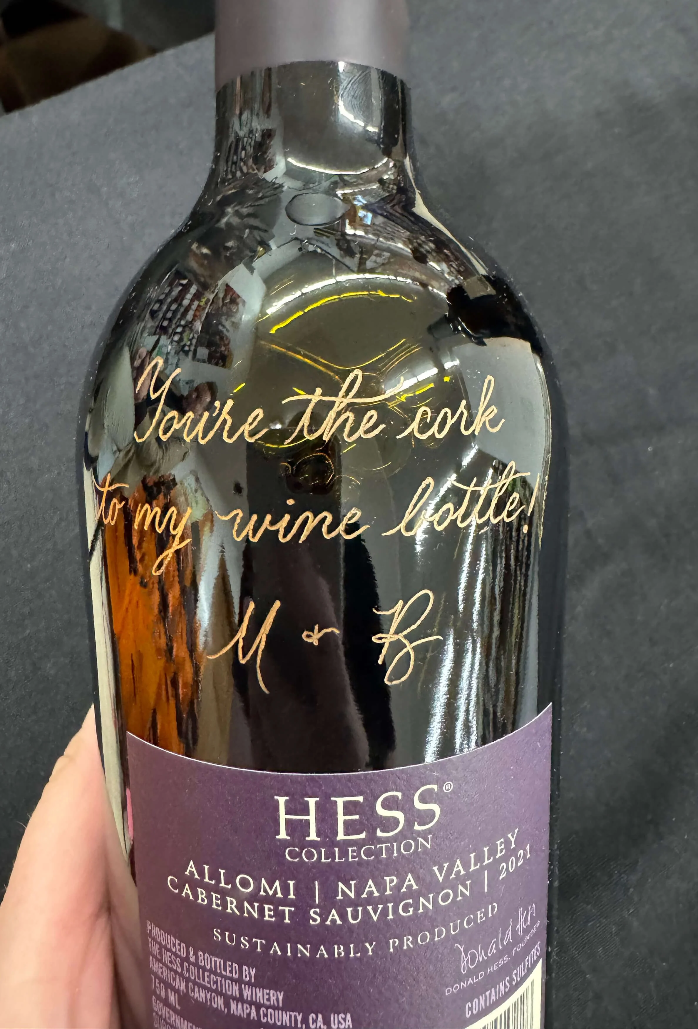 A wine bottle with a reflective surface and gold cursive text that reads, "You're the cork to my wine bottle!" The label indicates it is from the Hess Collection, Allomi Napa Valley Cabernet Sauvignon 2021, sustainably produced.