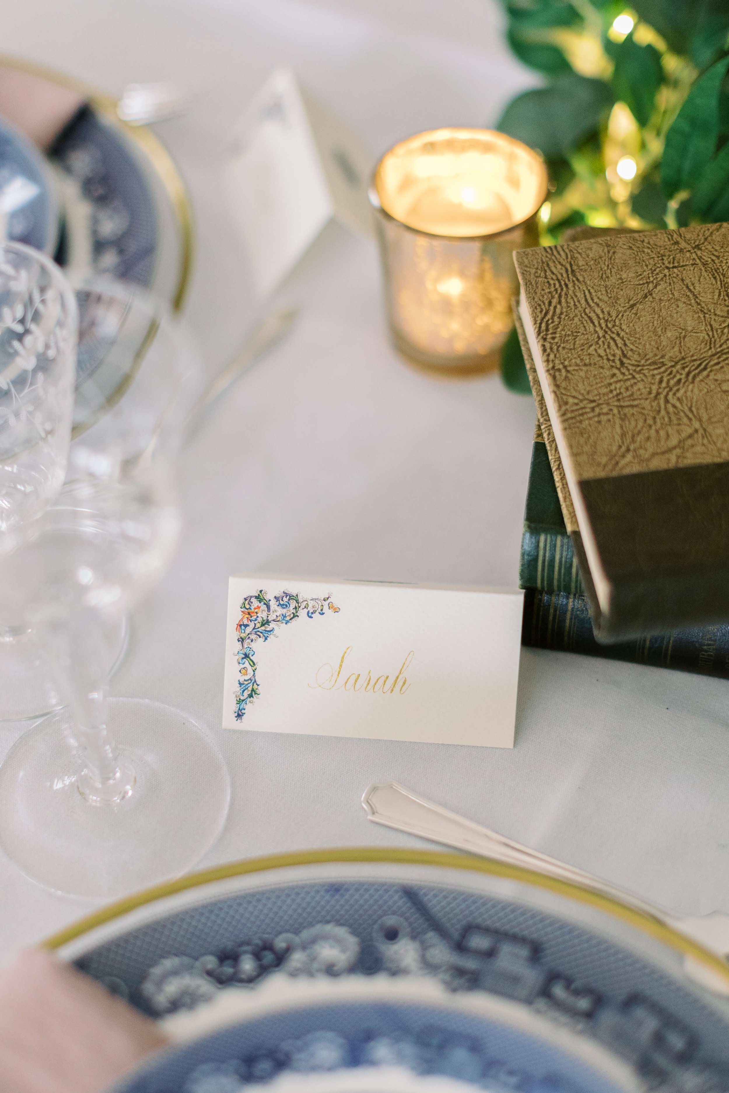 Place card with the name Sarah on a formal dining table, surrounded by wine glasses, books, and a lit candle.