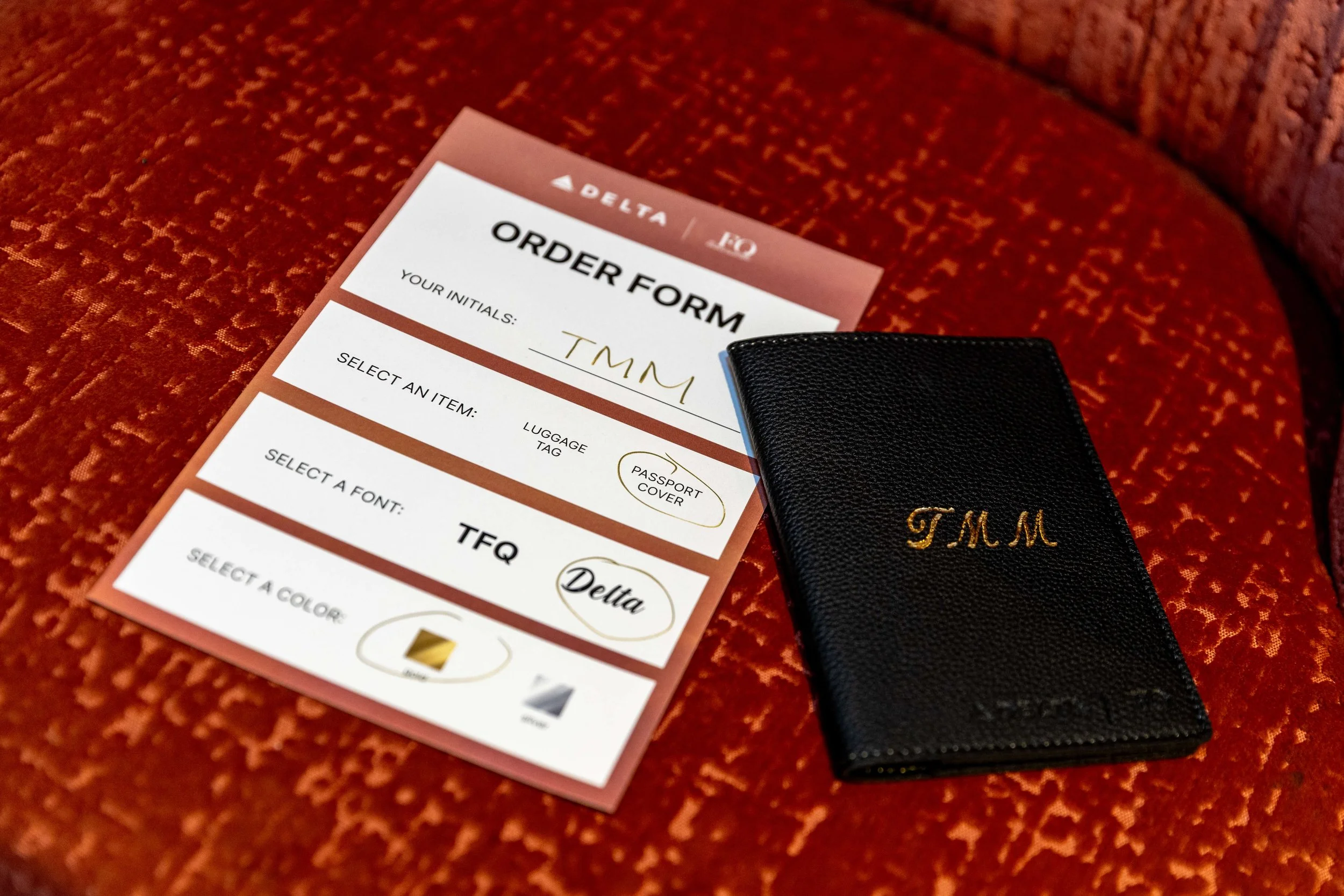 An order form, a black leather luggage tag with gold initials 'TMM', and a small black leather wallet on a textured red surface.