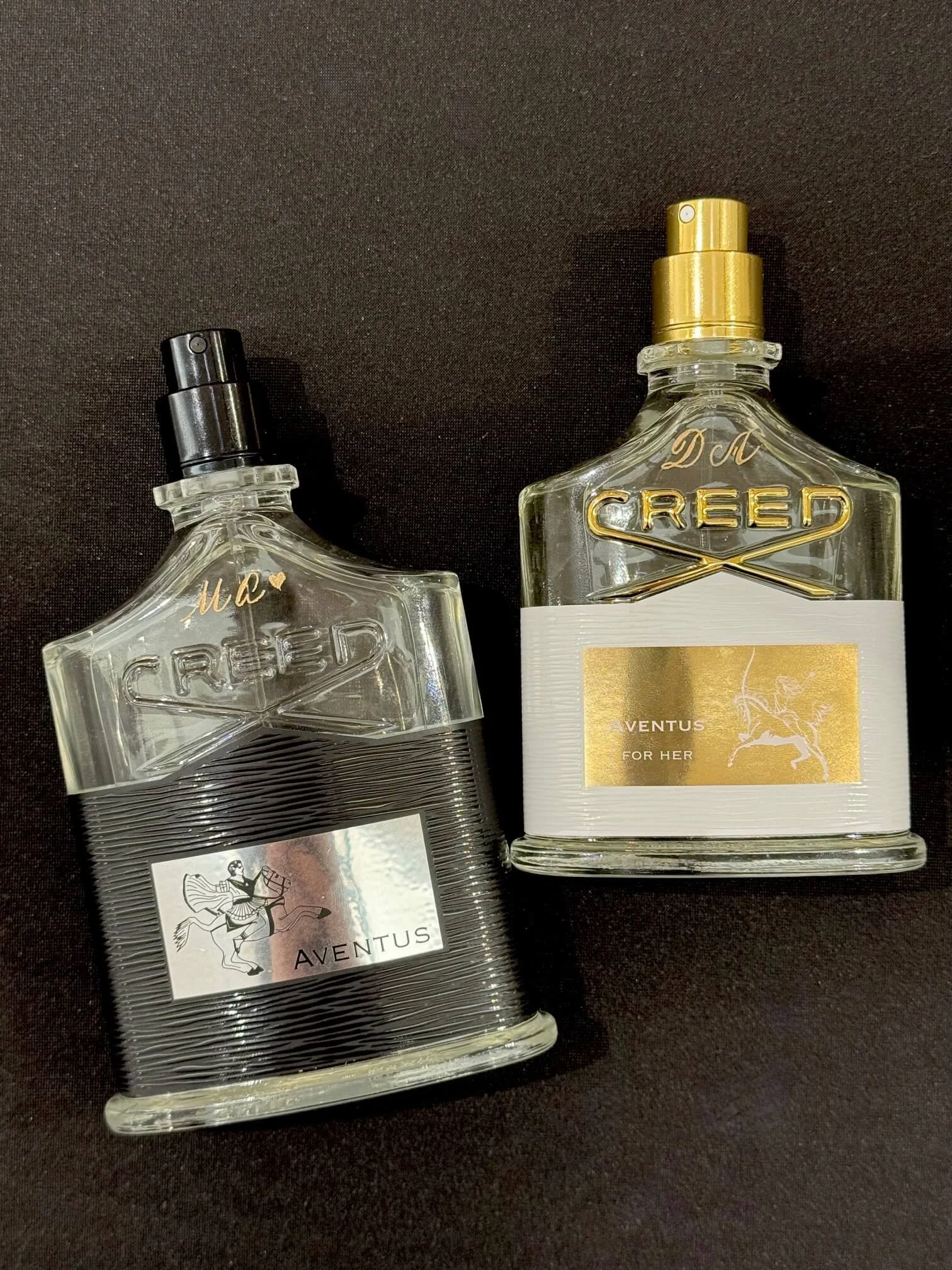 Two perfume bottles, one with a gold cap and the other with a black cap, on a dark surface.