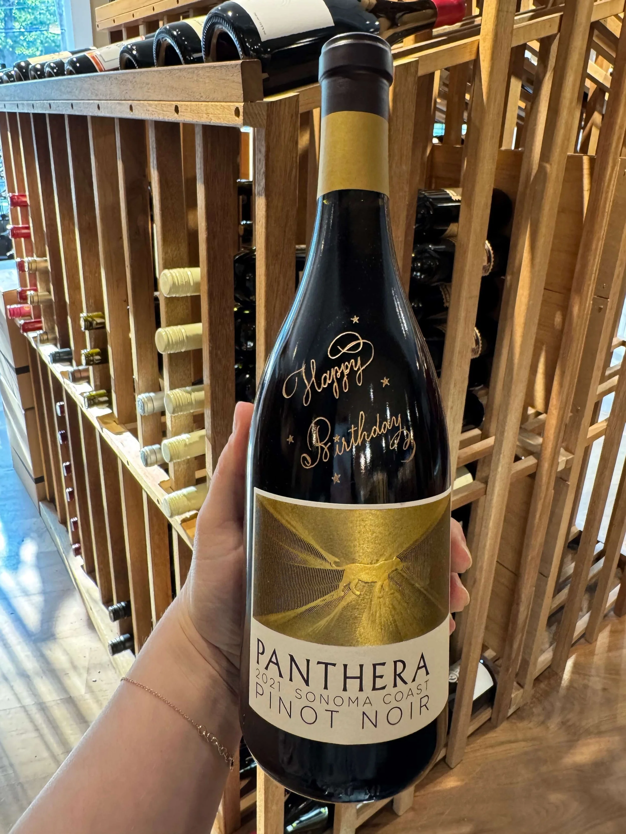 A person holding a bottle of 2021 Pantera Pinot Noir wine from Sonoma Coast, with a decorative gold label and a handwritten 'Happy Birthday' message on the bottle in a wine store.