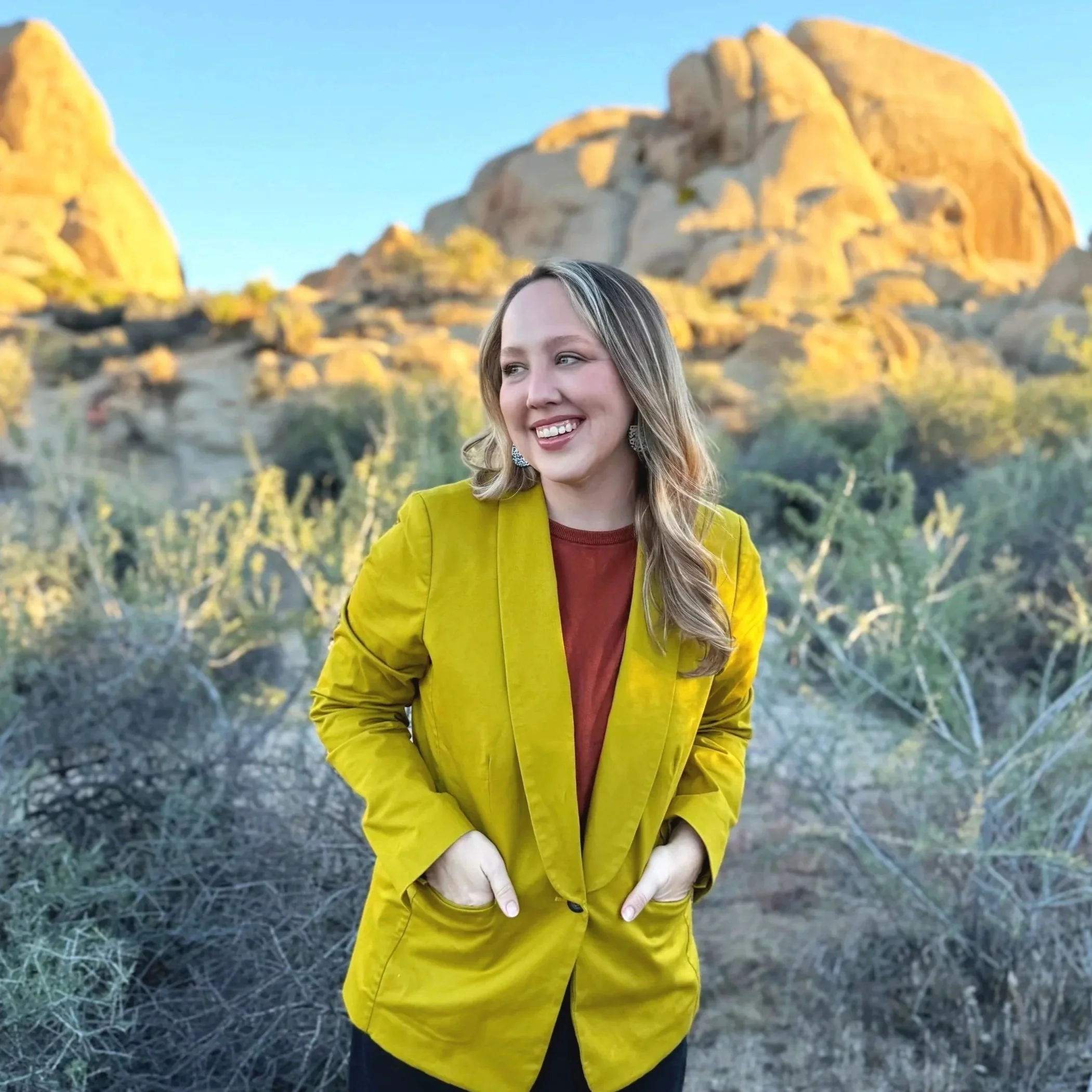 A woman in a yellow blazer and red top standing outdoors in a desert landscape with large rocks and sparse vegetation, smiling and looking to her right.