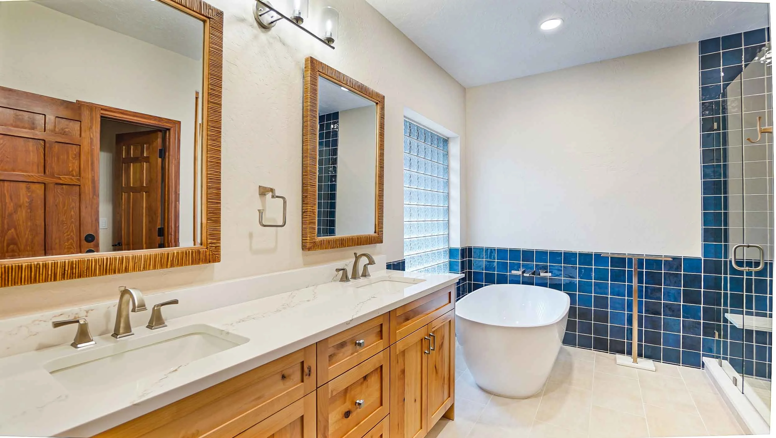 A modern bathroom built by home builders durango co featuring dual mirrors and sinks with a wooden vanity, a freestanding bathtub, blue tiled wall, glass block window, and a walk-in shower with a glass door.