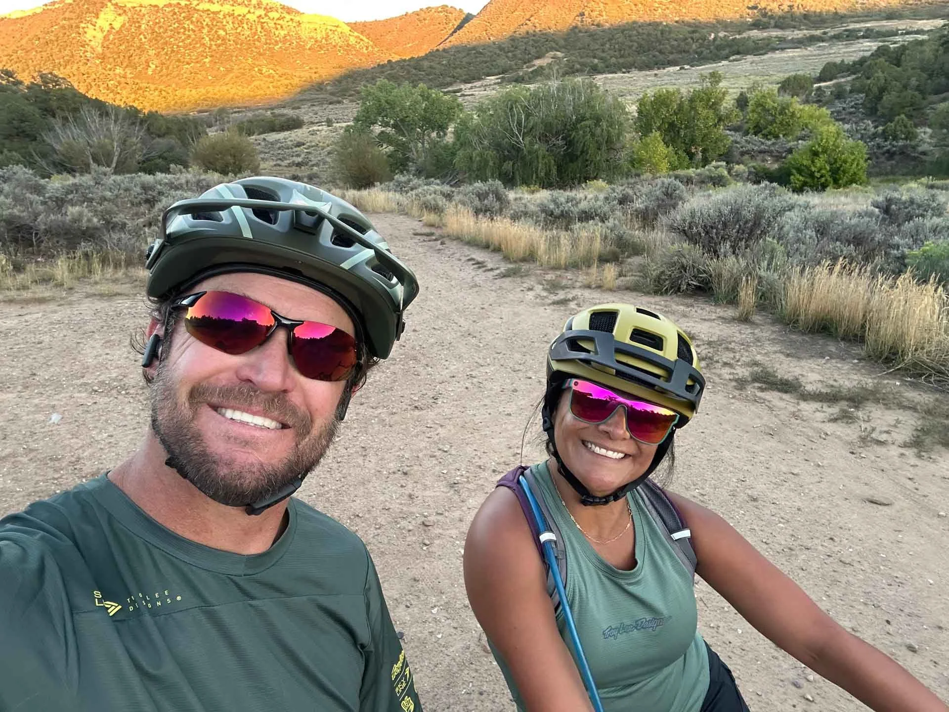 A man and woman wearing cycling helmets and sunglasses taking a selfie outdoors on a dirt trail surrounded by shrubbery and trees with hills in the background during sunset.