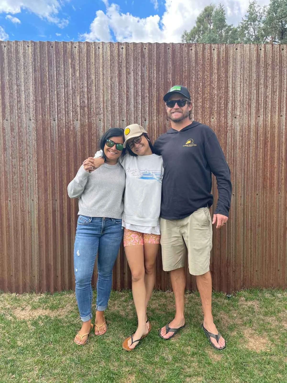 Three friends standing close together outdoors in front of a rusty fence, smiling and wearing casual summer clothes and sunglasses.