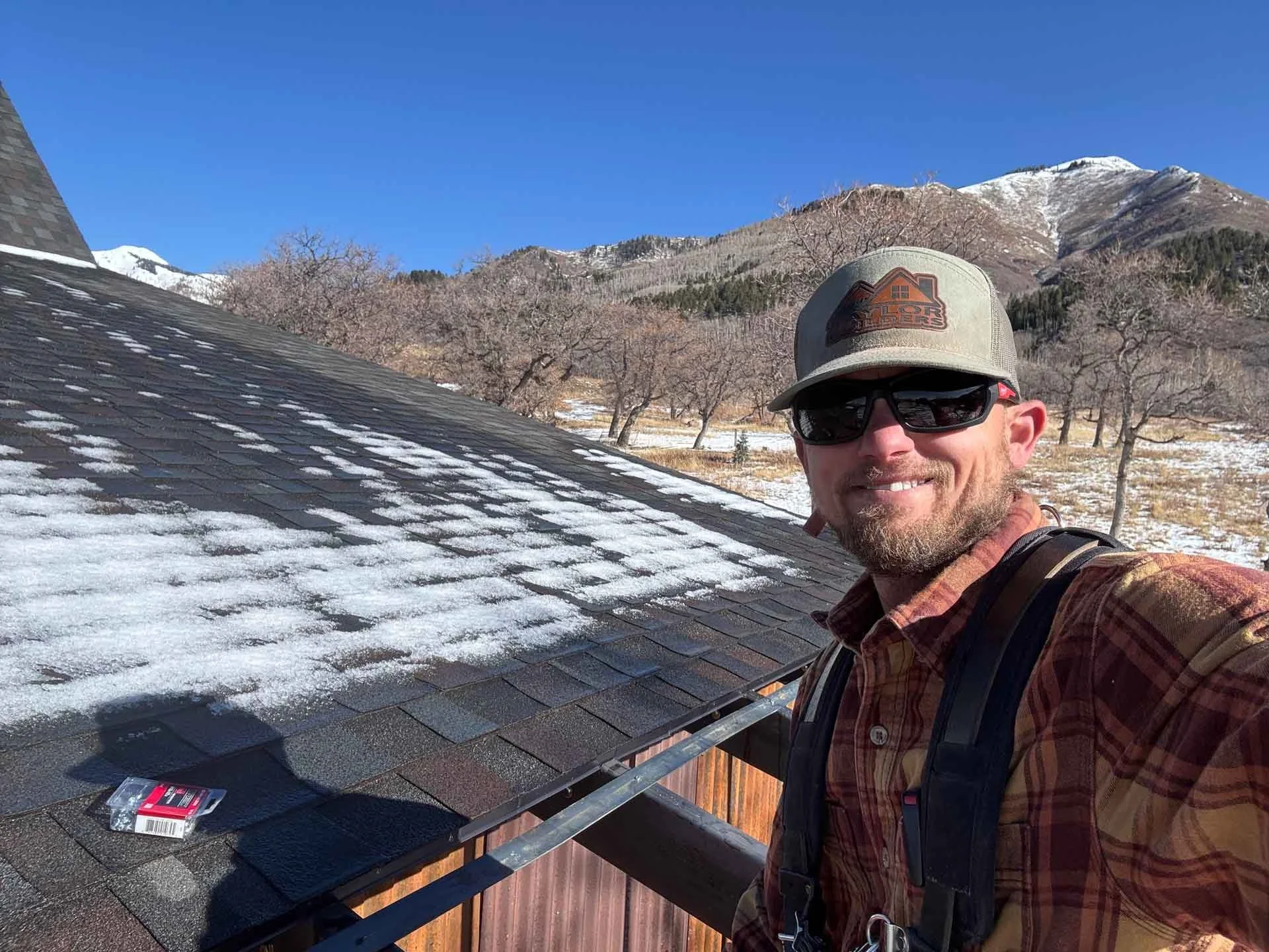 A man with sunglasses and a hat taking a selfie on a rooftop with snow patches. Background of snow-capped mountains, some trees, and a clear blue sky.