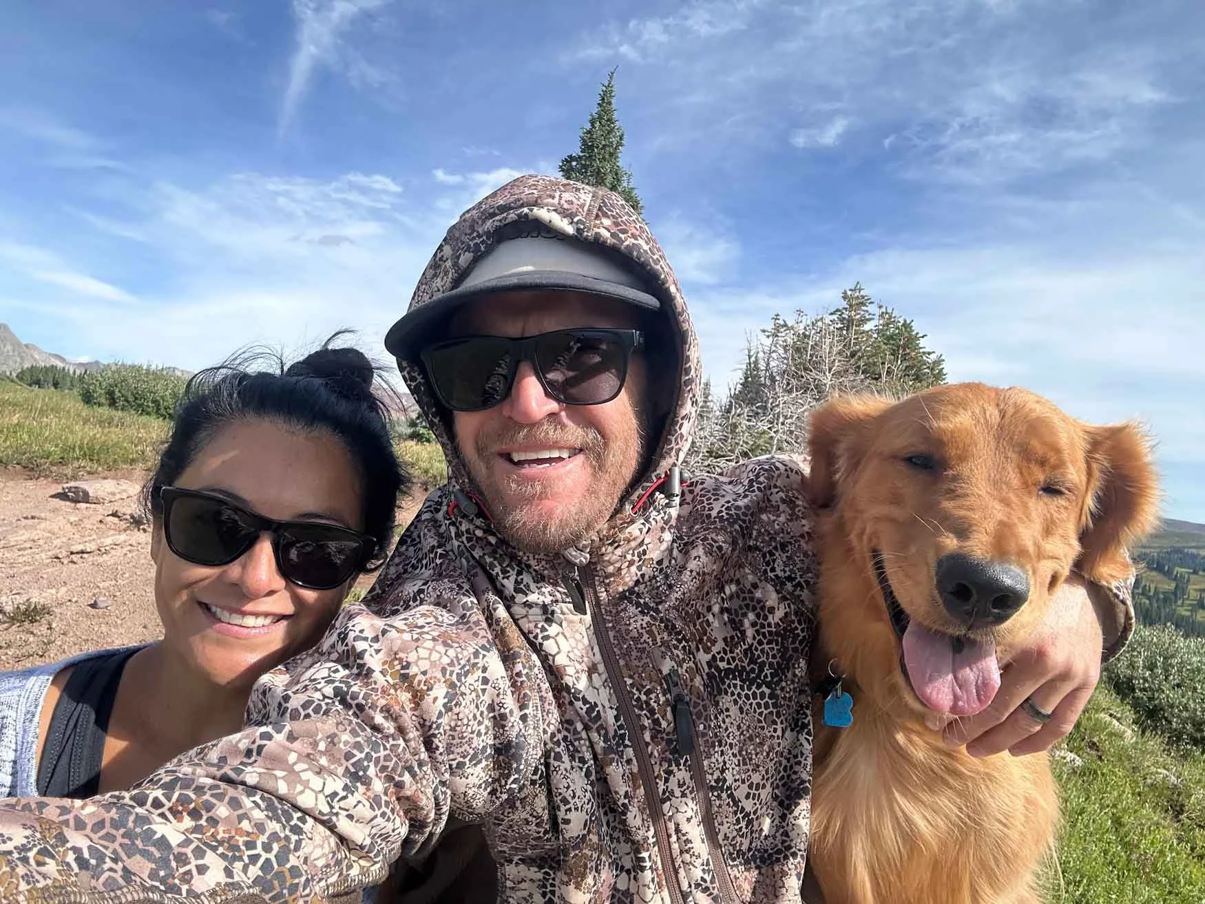 A smiling couple with black sunglasses taking a selfie outdoors on a sunny day, accompanied by a golden retriever dog with its tongue out, in a scenic landscape with green trees, mountains, and a blue sky with wispy clouds.