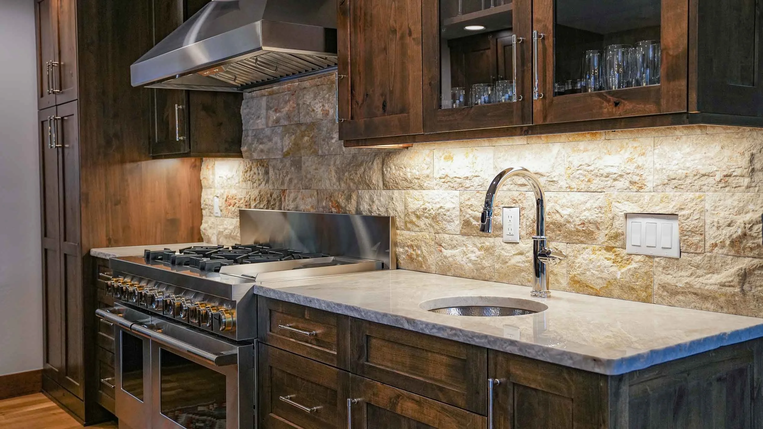 Kitchen with dark wood cabinets, a granite countertop, and a stone backsplash. Includes a stove with a vent hood and a faucet over a small sink built by remodeling contractors