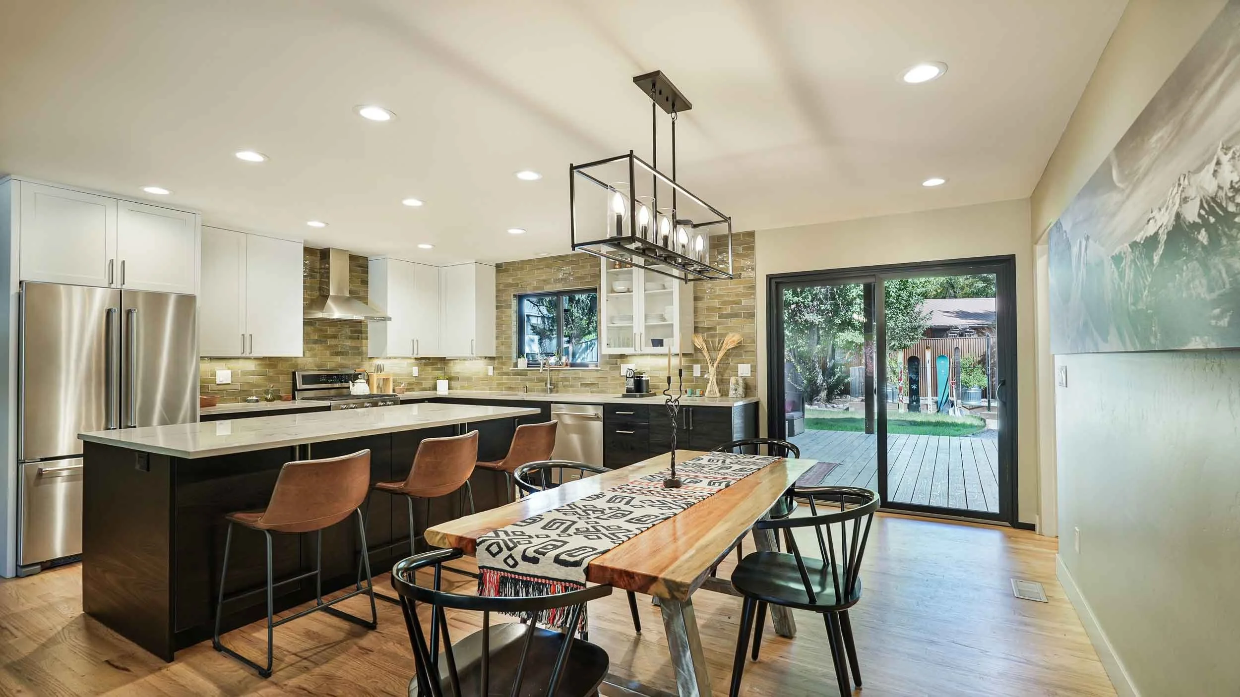 Open-concept kitchen and dining area with black and white cabinets, stainless steel appliances, a brick backsplash, and a wooden dining table with black and brown chairs, overlooking a backyard with trees and a deck.