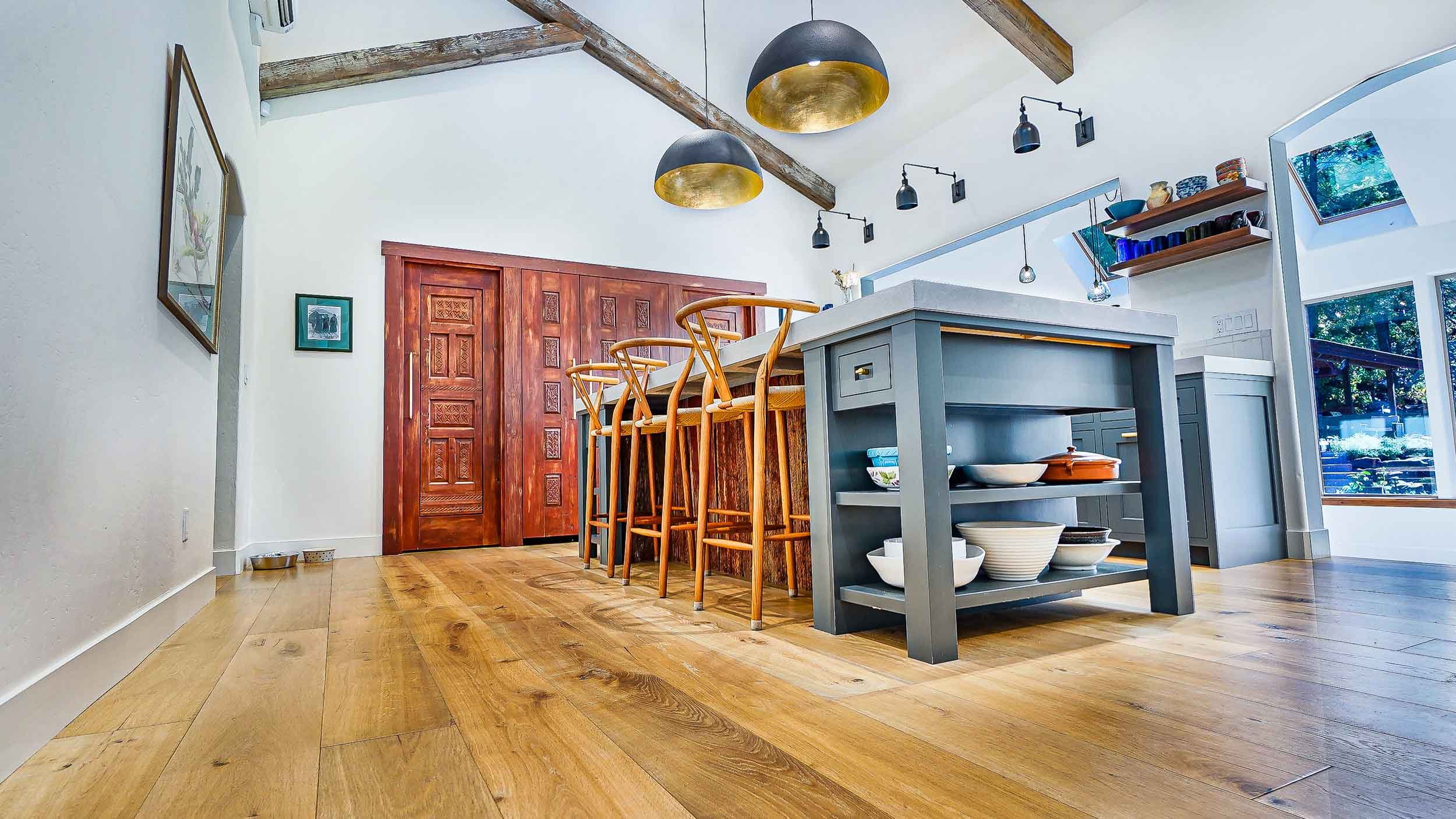 Modern kitchen with wooden floor, gray island, wooden bar stools, artwork on white walls, wood sliding barn door, and large window with views of trees