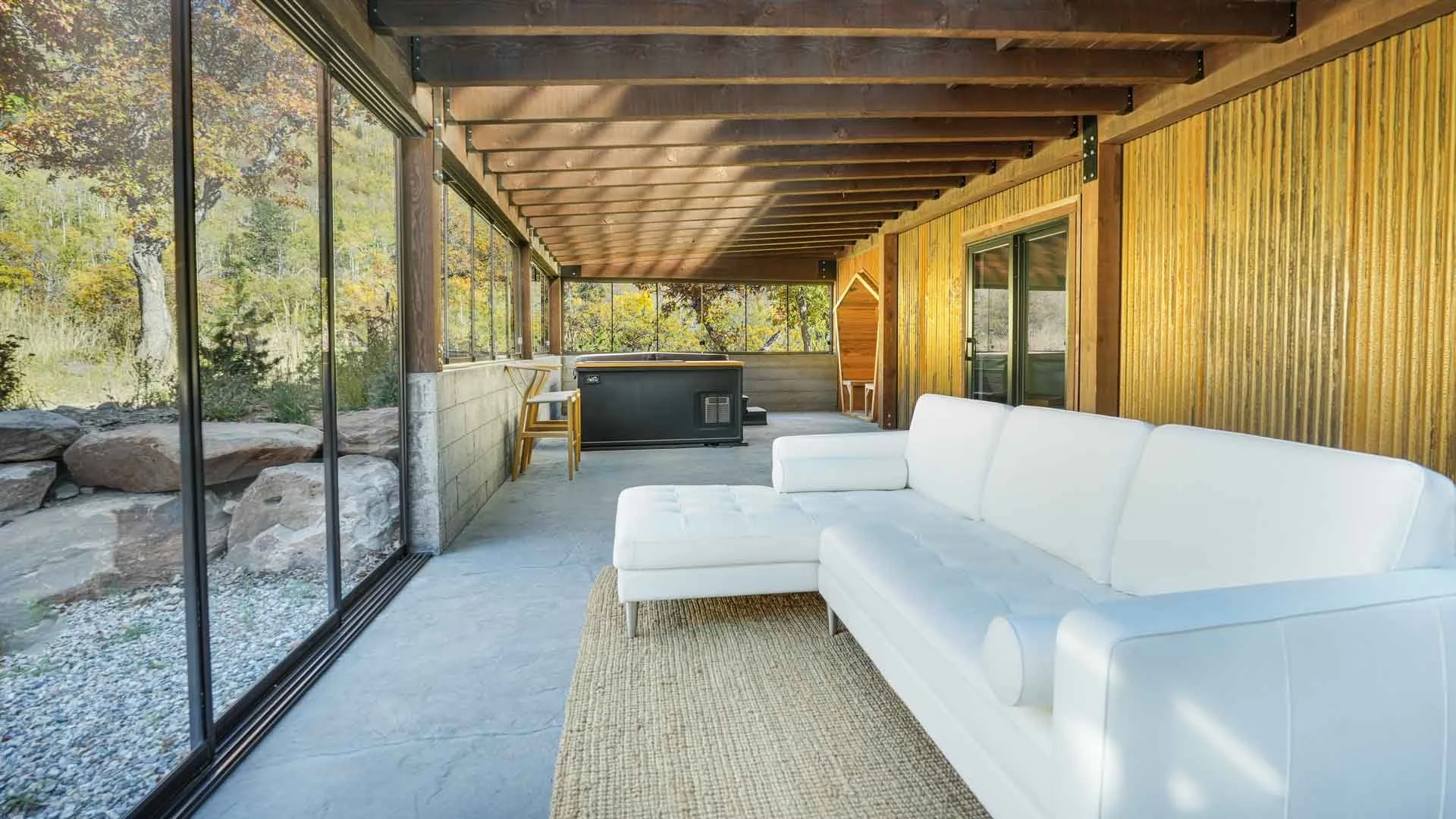 Modern indoor porch with large glass sliding doors, white sectional sofa, beige rug, hot tub, wooden ceiling beams, and view of trees outside built by custom home builders durango