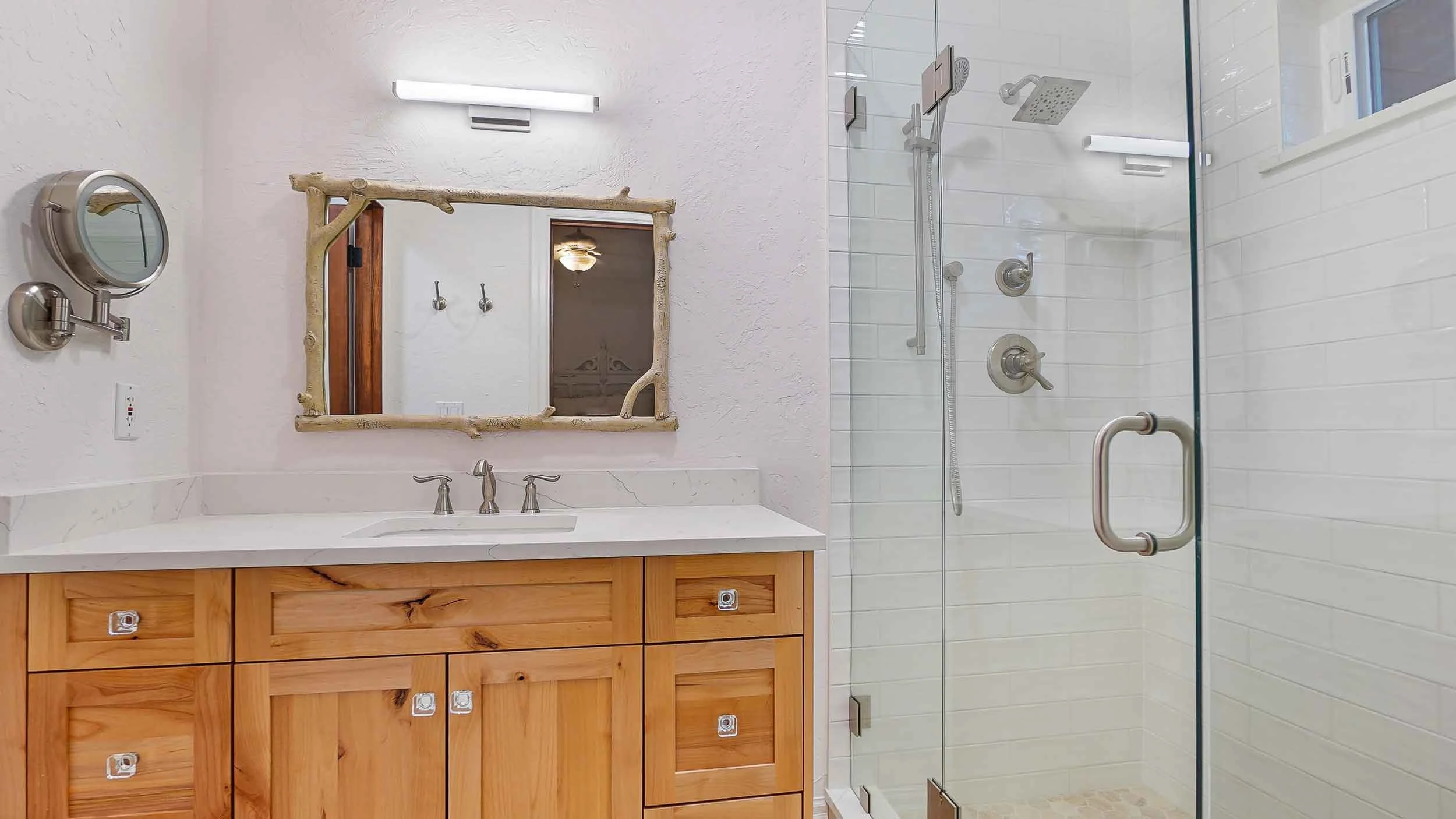Bathroom with wooden vanity, white countertop, oval mirror, and walk-in shower with glass door, white tiles, and multiple showerheads built by home contractors durango
