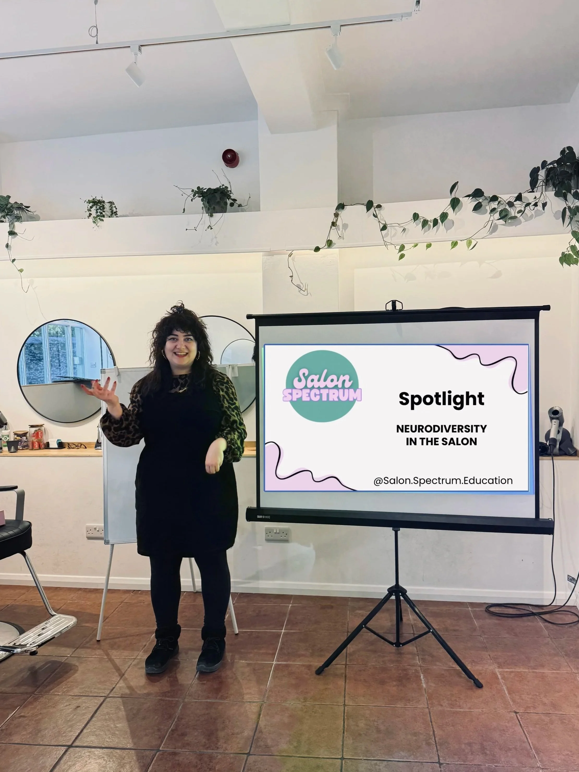 Educator presenting a ‘Neurodiversity in the Salon’ training session at Salon Spectrum, teaching hairstylists how to create inclusive. The woman is smiling, wearing a black outfit and leopard print sleeves, in a room with mirrors, plants, and chairs.