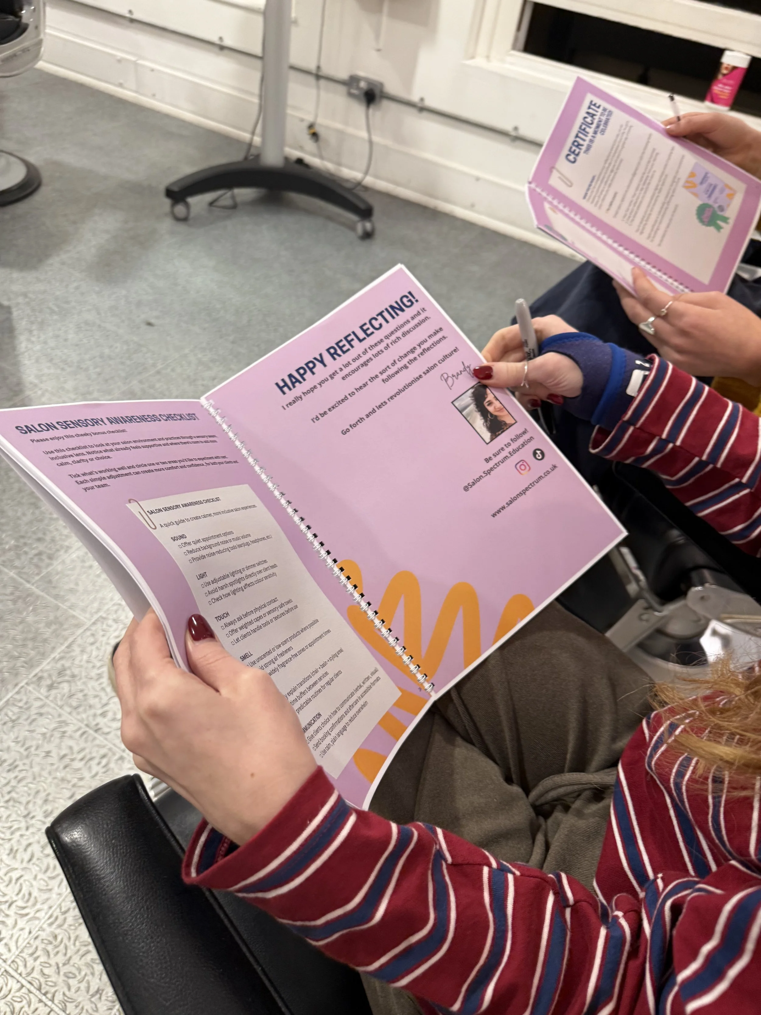 Hairdresser reading the Salon Spectrum Reflective Workbook in a salon setting, with another person holding a certificate in the background.