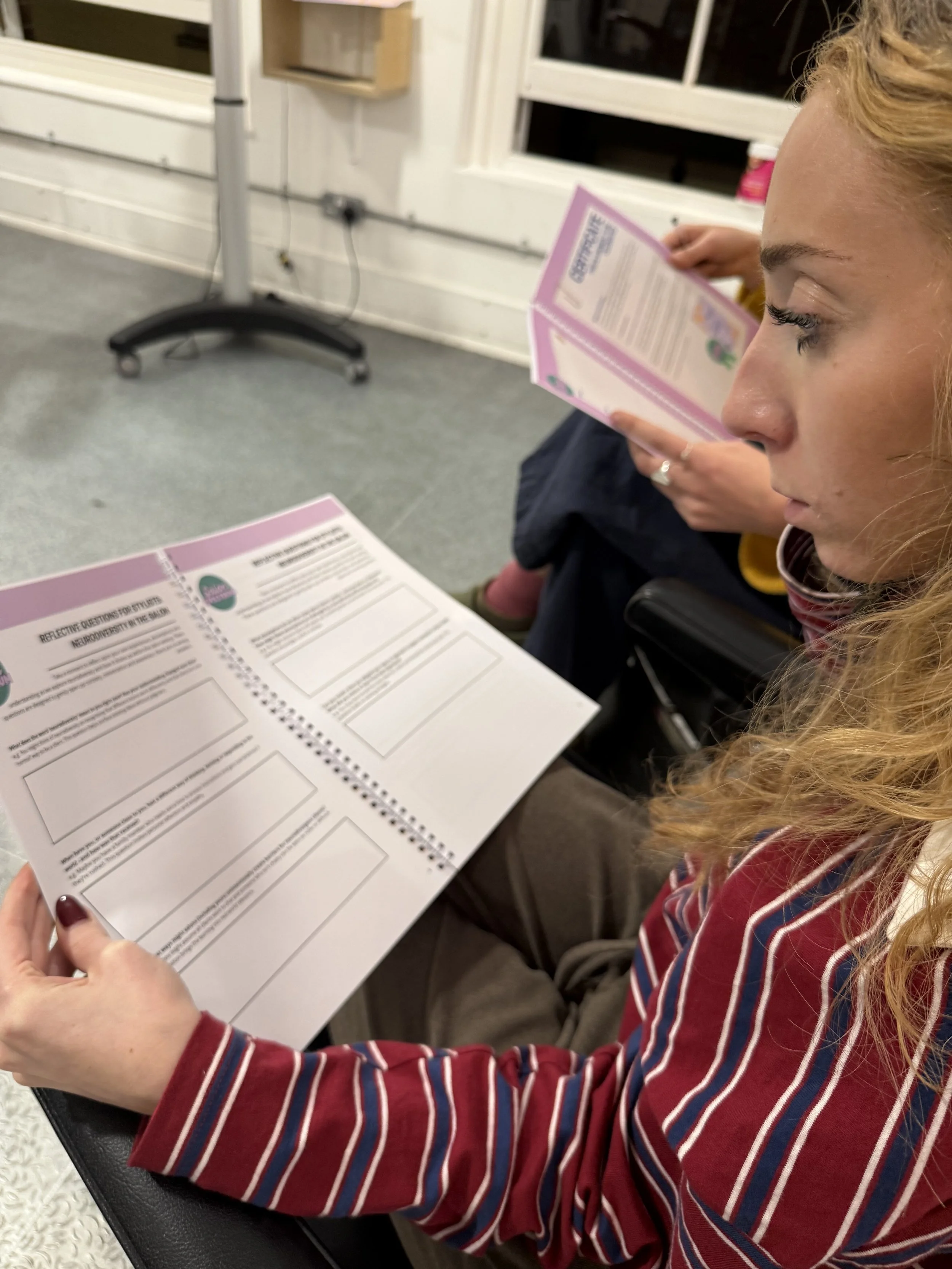 A hairdresser with curly blonde hair, sitting in the salon looking at the Reflective Workbook for stylists. The workbook has text and blank boxes. She is sitting indoors on a red sofa in the salon.