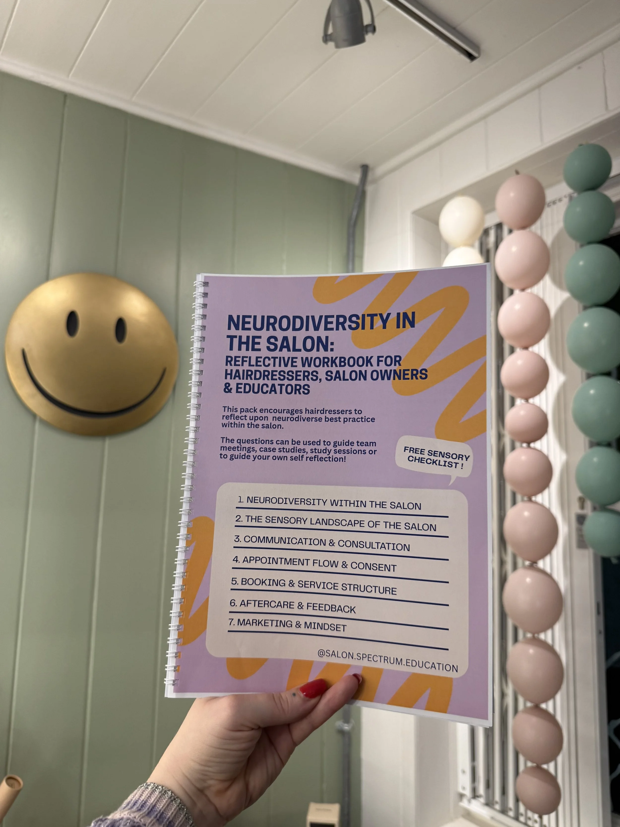 A person holding a purple booklet titled "Neurodiversity in the Salon: Reflective Workbook for Hairdressers, Salon Owners & Educators" in a salon with pastel-colored decorations, including a yellow smiley face wall hanging and a string of pastel beads.