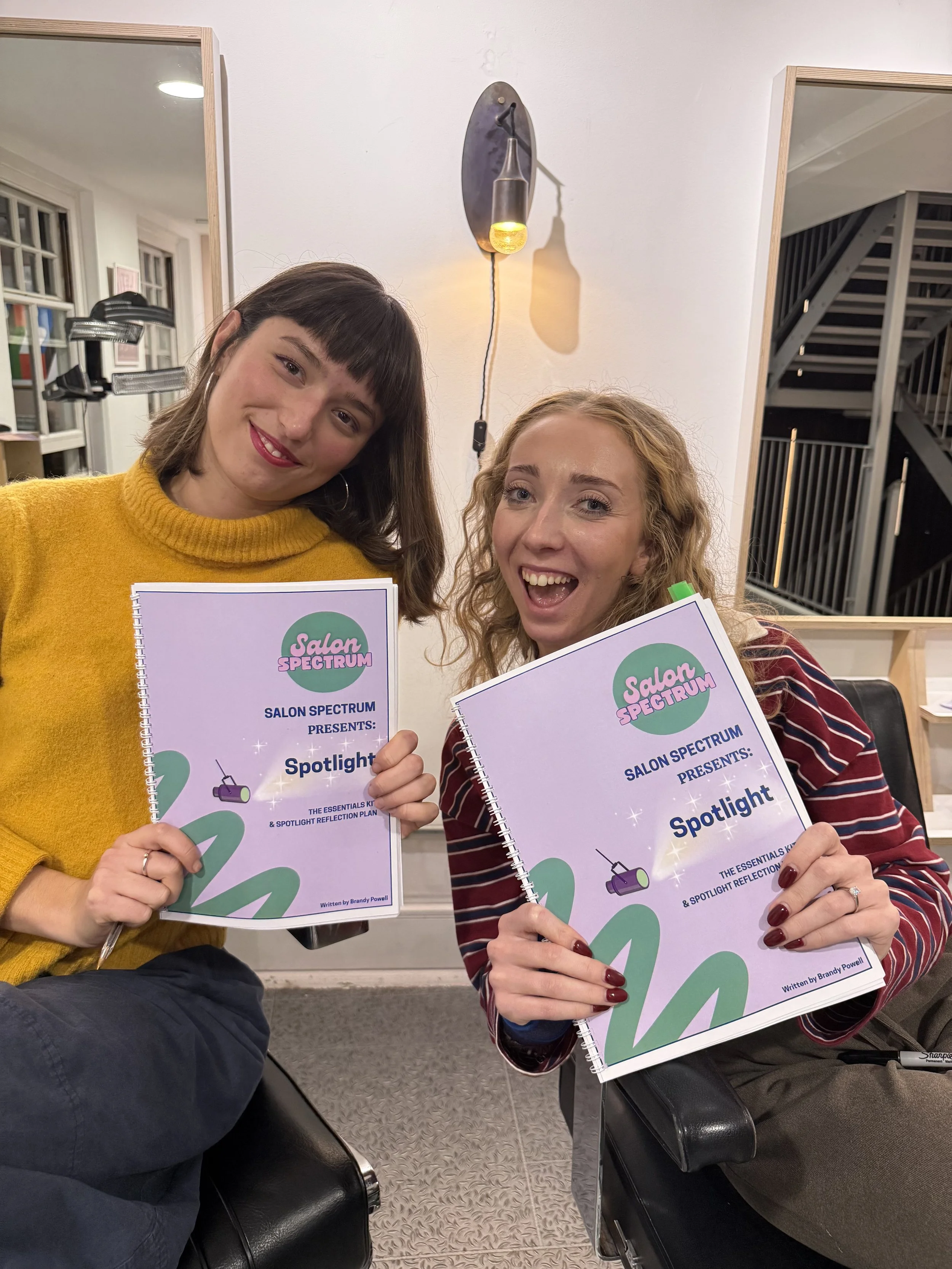 Two hairstylists smiling and holding their Salon Spectrum ‘Spotlight’ neurodiversity-in-the-salon workbooks during an inclusive education workshop.