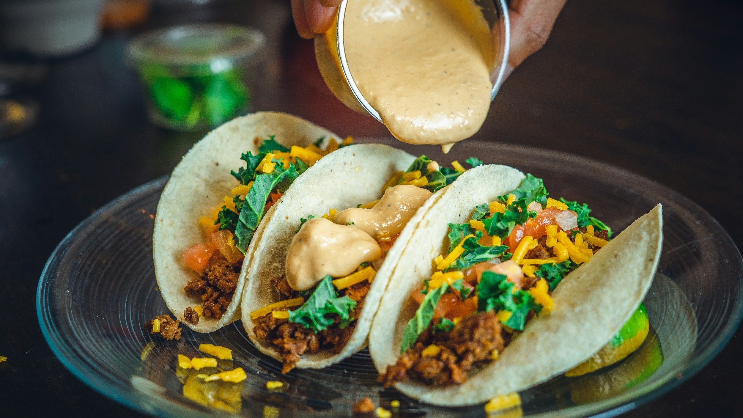Close-up of three tacos filled with ground beef, lettuce, shredded cheddar cheese, diced tomatoes, and topped with creamy sauce, all arranged on a glass plate.