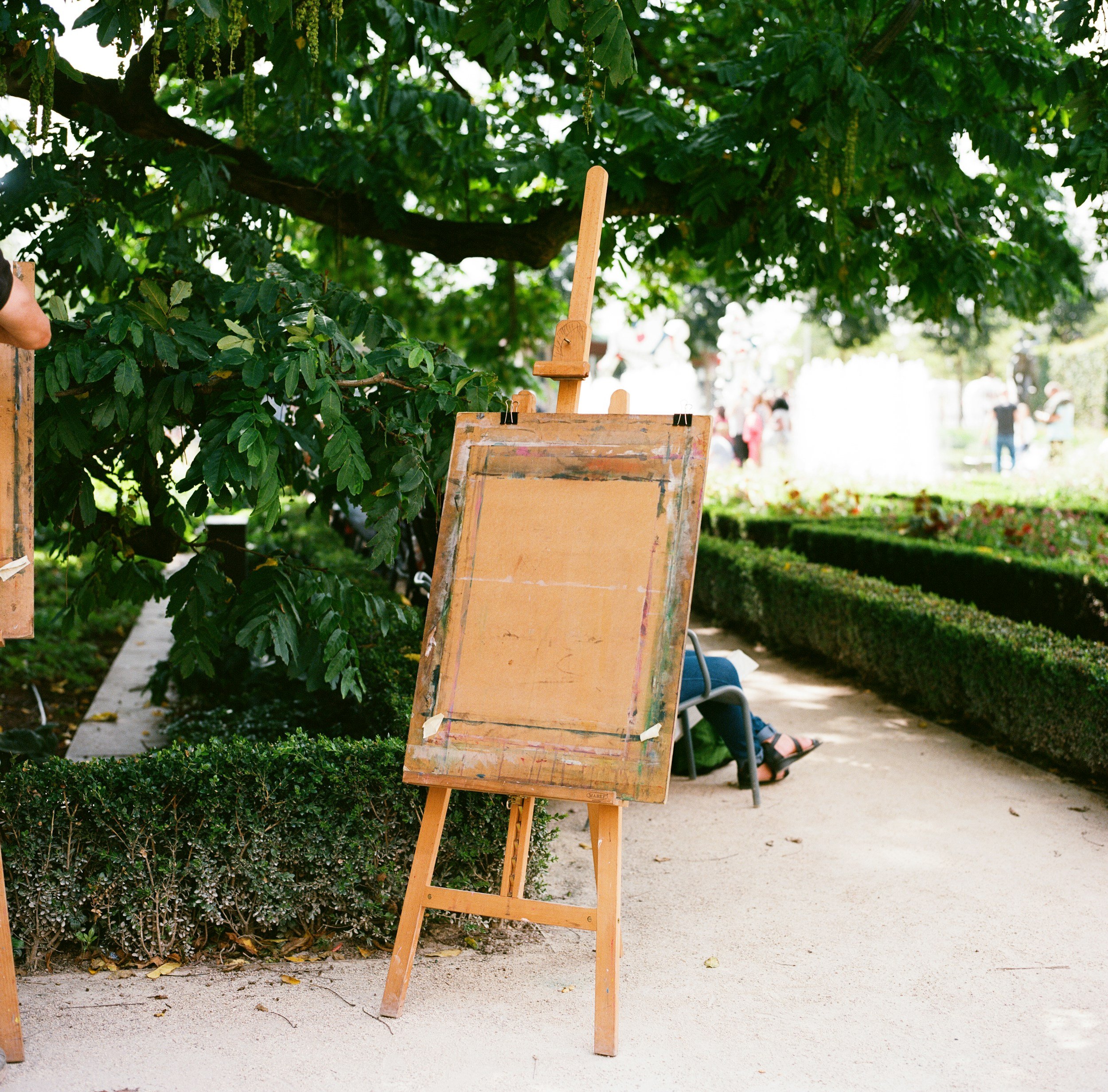Empty wooden artist's easel standing on a sidewalk beneath a leafy tree in a park.