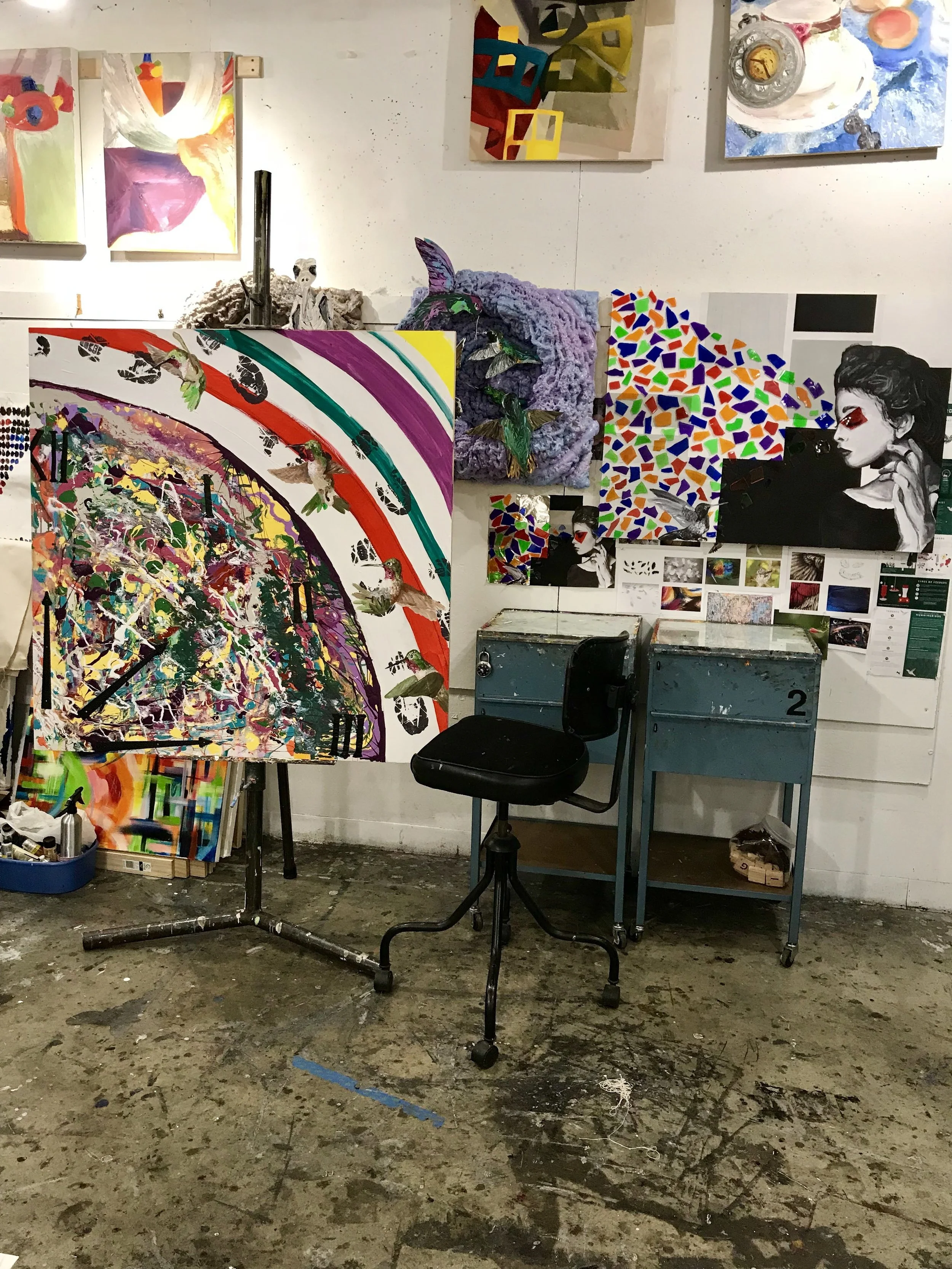 An art studio with various colorful paintings on the wall and on easels, a chair, and a cluttered floor with paint splatters.