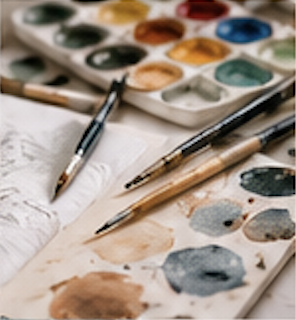 Artist's watercolor palette with various colors, watercolor brushes, and a paintbrush on paper.