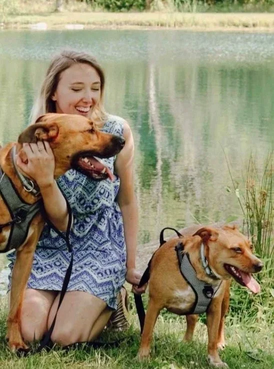 A woman kneeling beside a lake with two large dogs, all appearing happy and relaxed. The woman is smiling while petting one dog and the other dog stands nearby.
