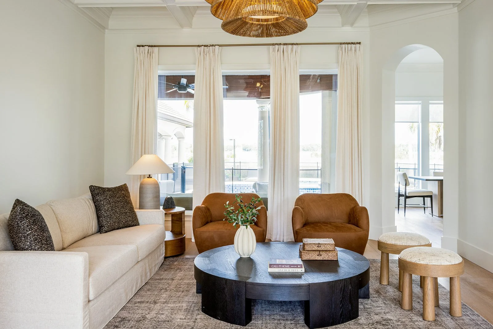 Living room with a beige sofa, two brown armchairs, a black round coffee table with a white vase and books, beige side table with a lamp, two small beige stools, and large windows with white curtains showing an outdoor patio.